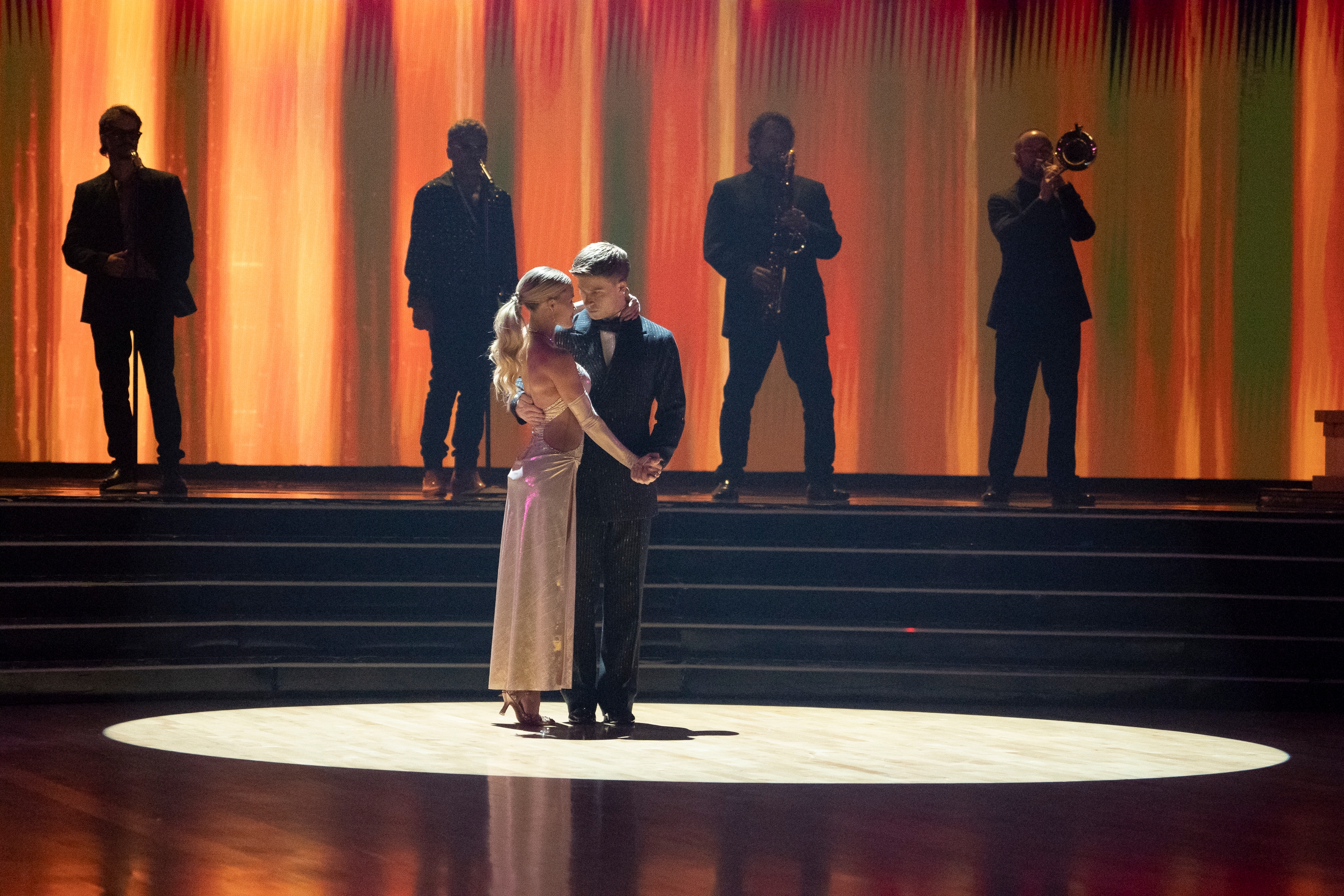 Couple in formal dance attire embrace on stage, silhouetted musicians play in the background
