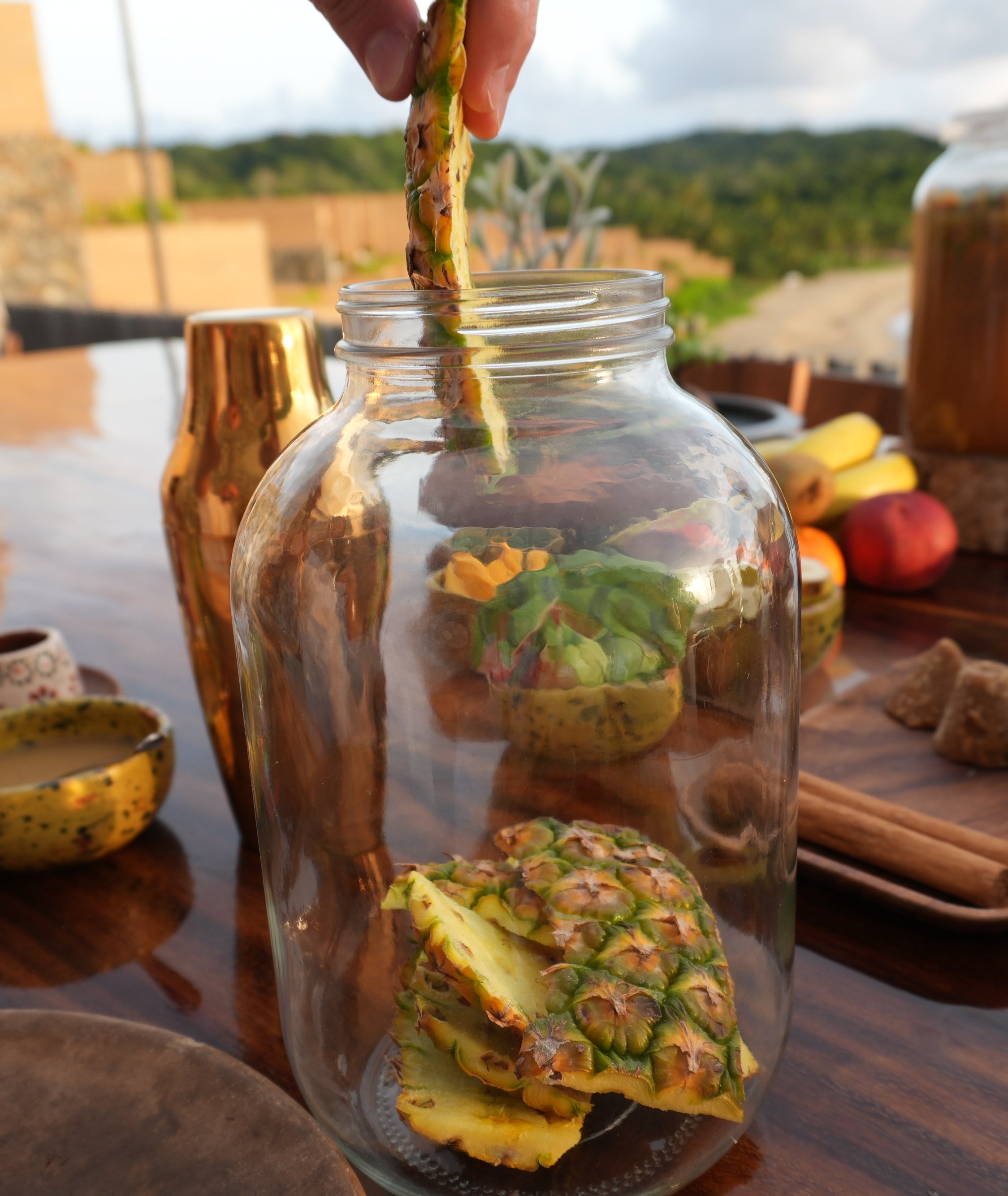 Hand placing pineapple pieces into a large jar on an outdoor table with various spices and ingredients around. Scenic view in the background