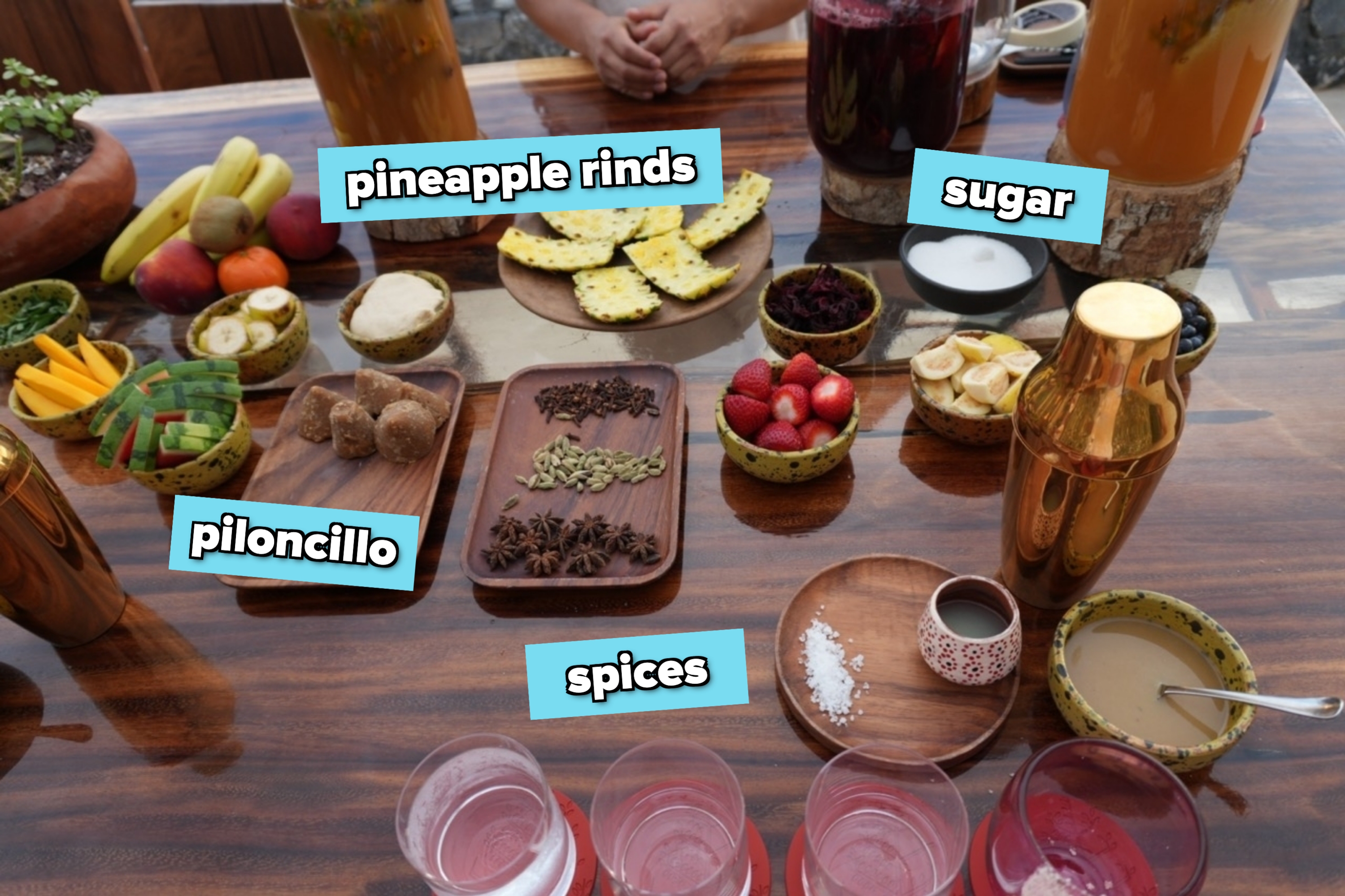 A wooden table with various fresh fruits, spices, drinks, and a pair of hands resting on it, suggesting a setting for juicing or food preparation