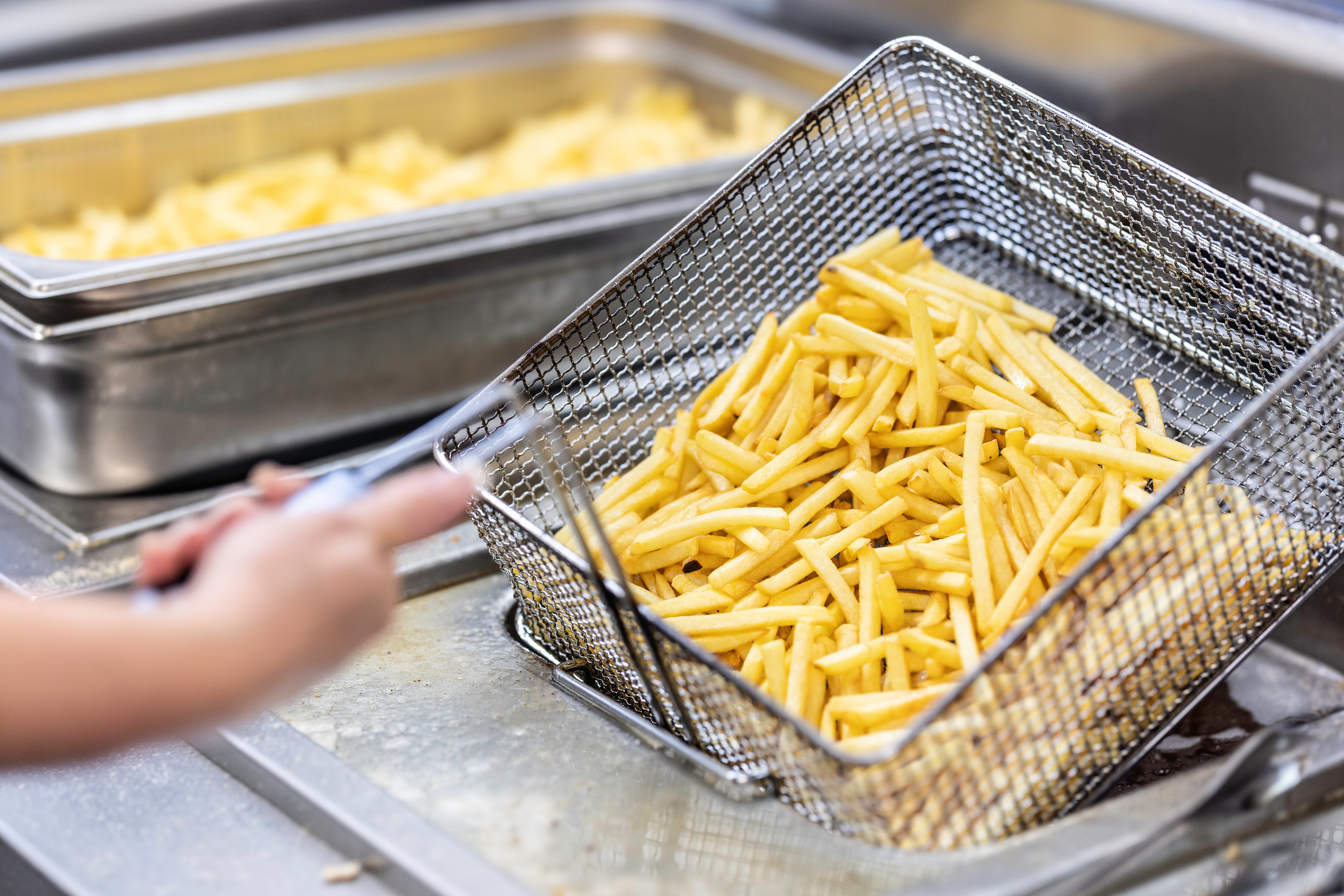 A person lifts a metal fryer basket filled with freshly cooked French fries from a deep fryer
