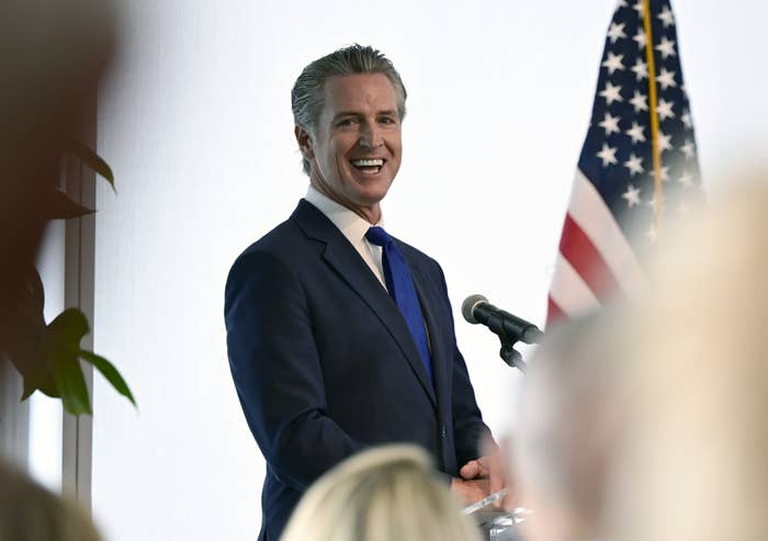 Gavin Newsom speaking at a podium with an American flag in the background, smiling at an audience
