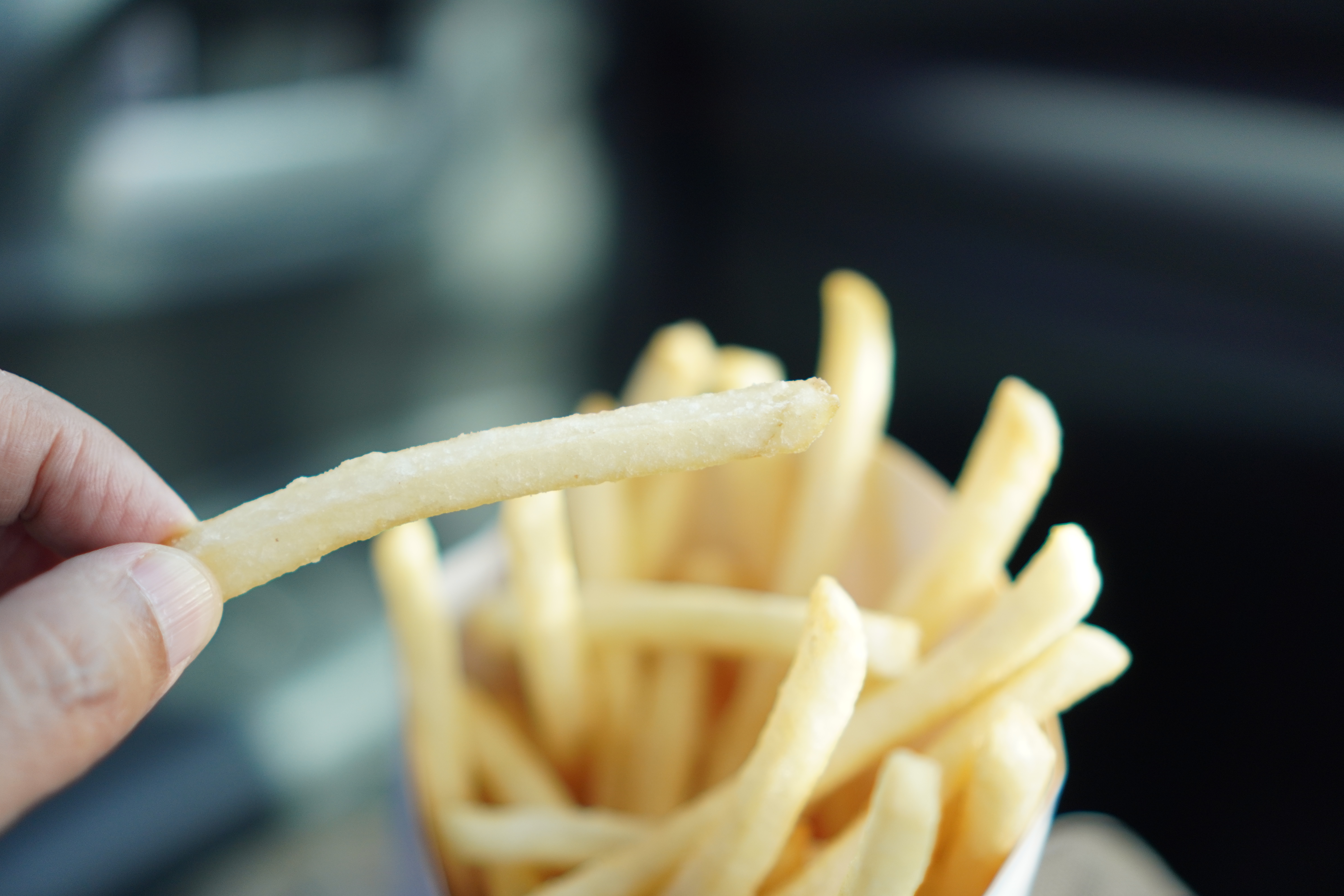 A hand holds a single French fry above a container filled with more fries