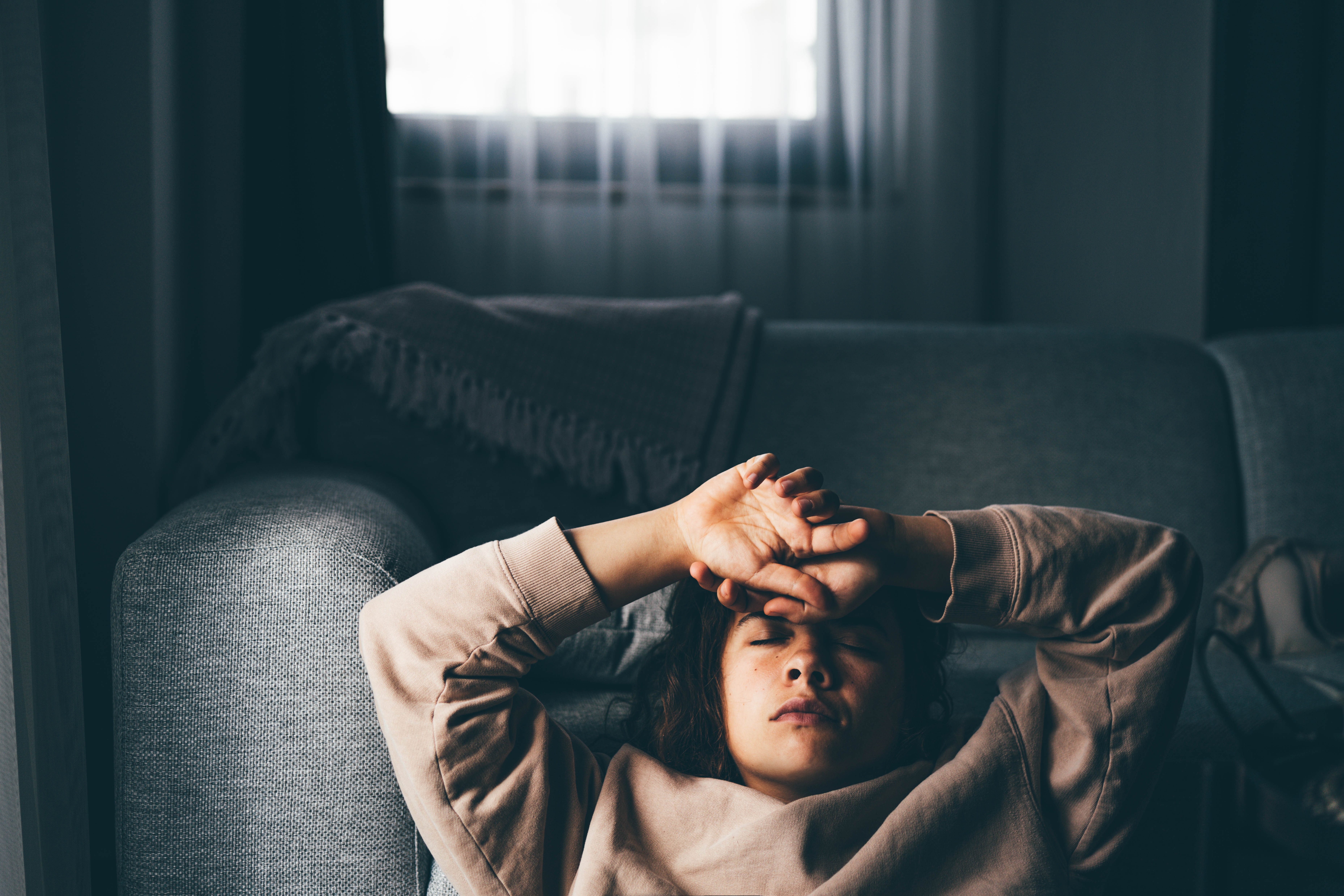 Personen som ligger på en sofa, lukkede øyne, armene hviler på pannen, virker avslappet og avslappende. Koselig hjemmemiljø i bakgrunnen