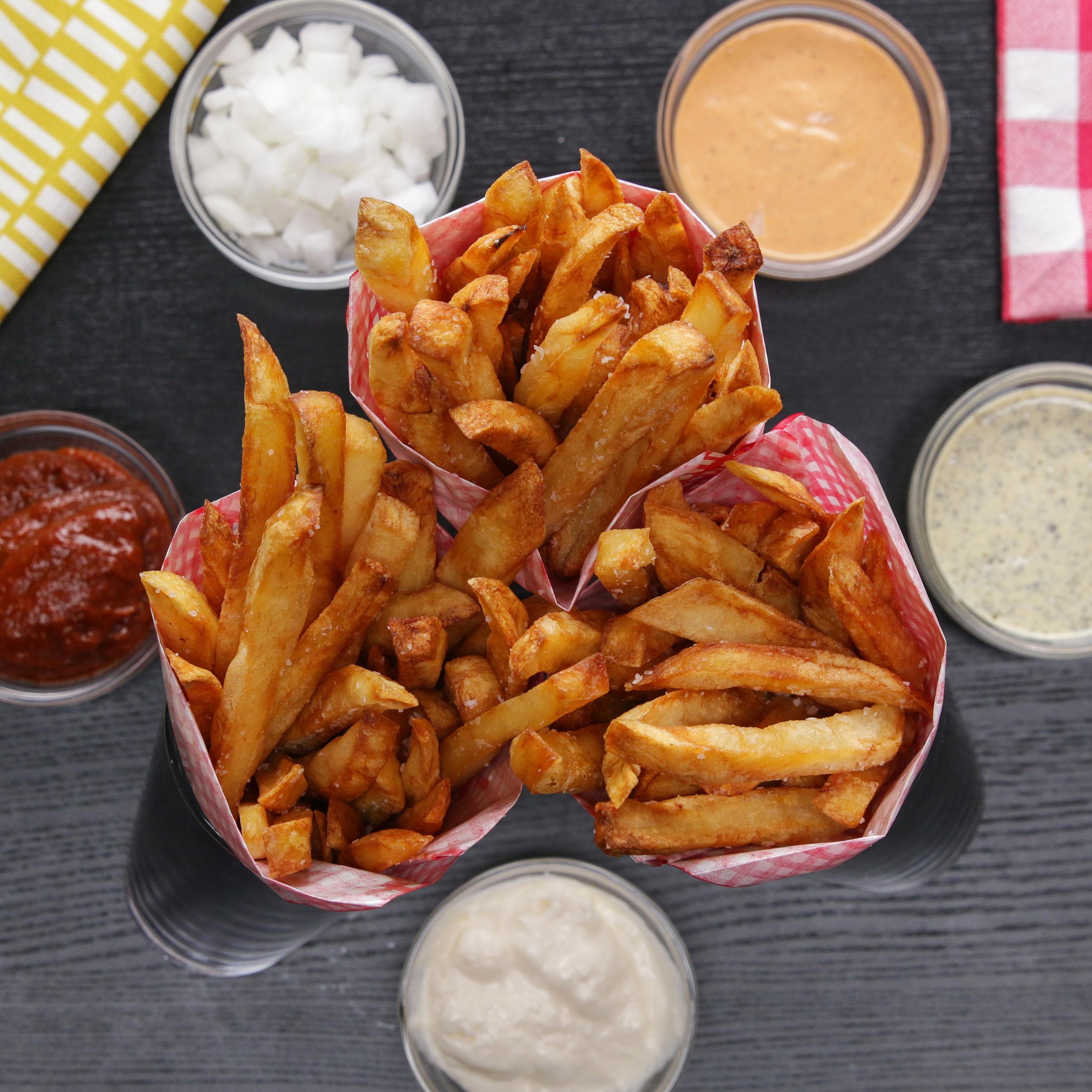Fries in paper cones surrounded by dipping sauces, including ketchup, mayonnaise, and mustard, on a dark surface