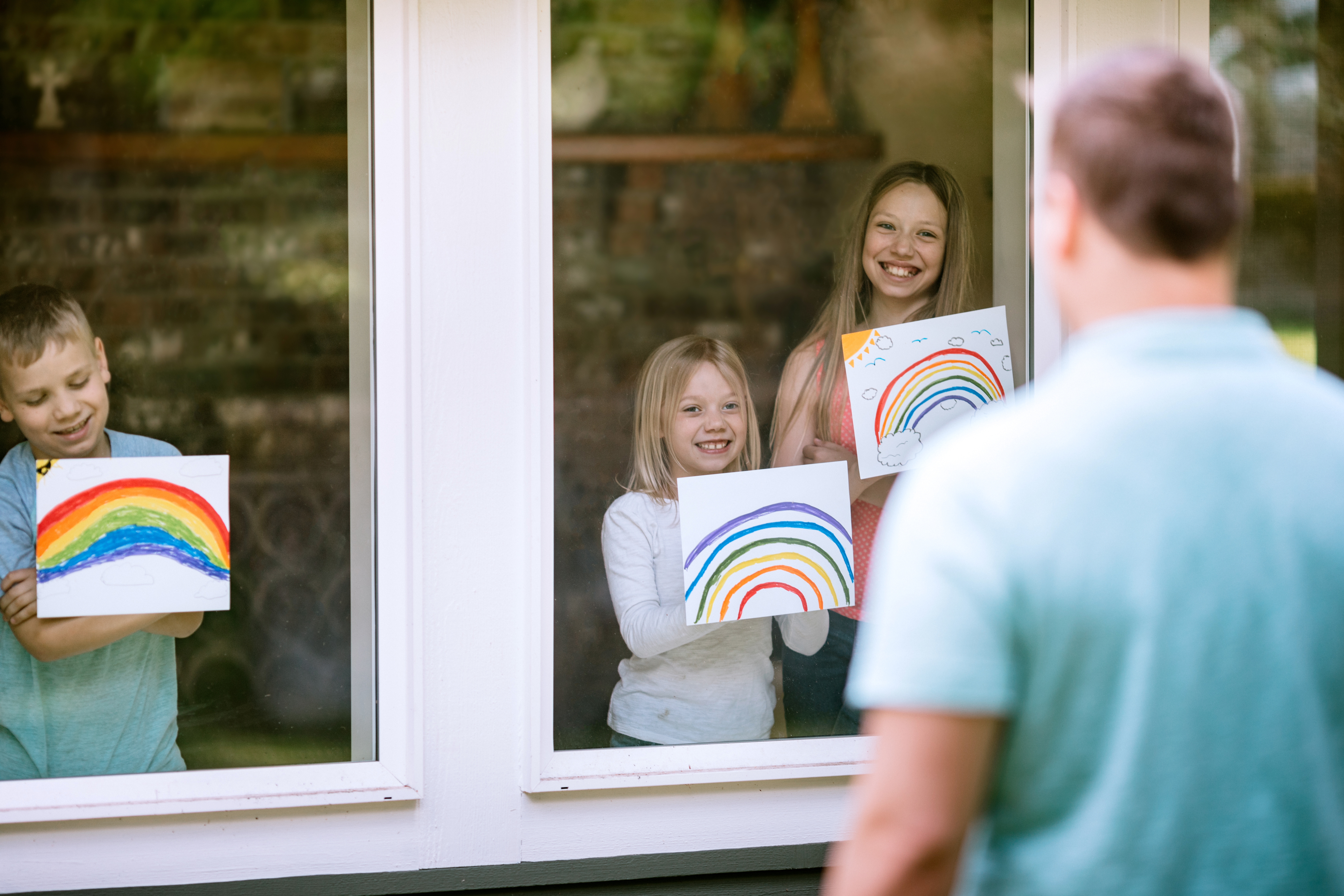Tre barn inne i et hjem viser regnbuens tegninger gjennom et vindu til en mann som står utenfor, smiler og nyter kunsten sin