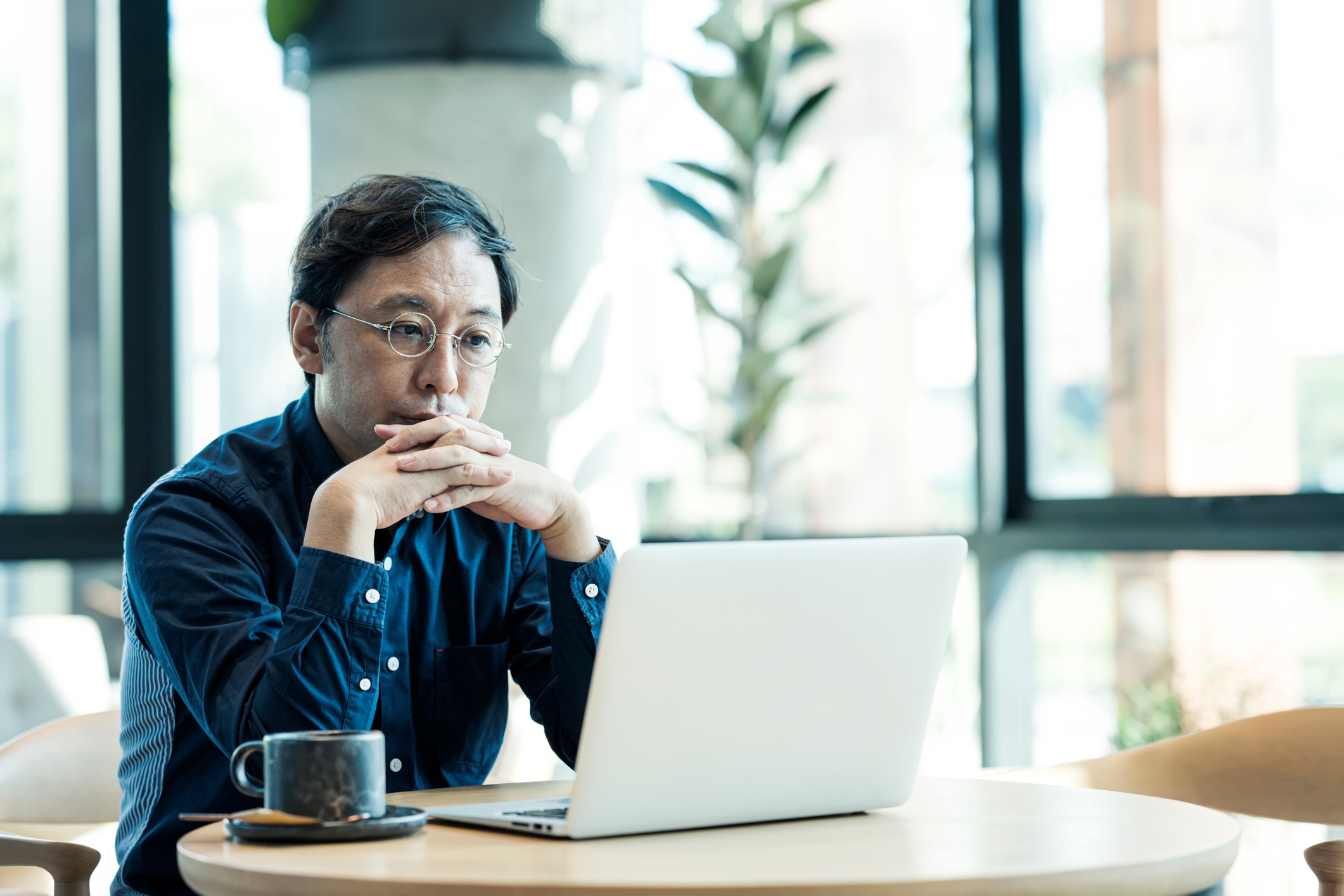 Person i briller og blå skjorte sitter ved et kafébord, og ser ettertenksomt på en bærbar datamaskin. En kaffekopp er ved siden av den bærbare datamaskinen