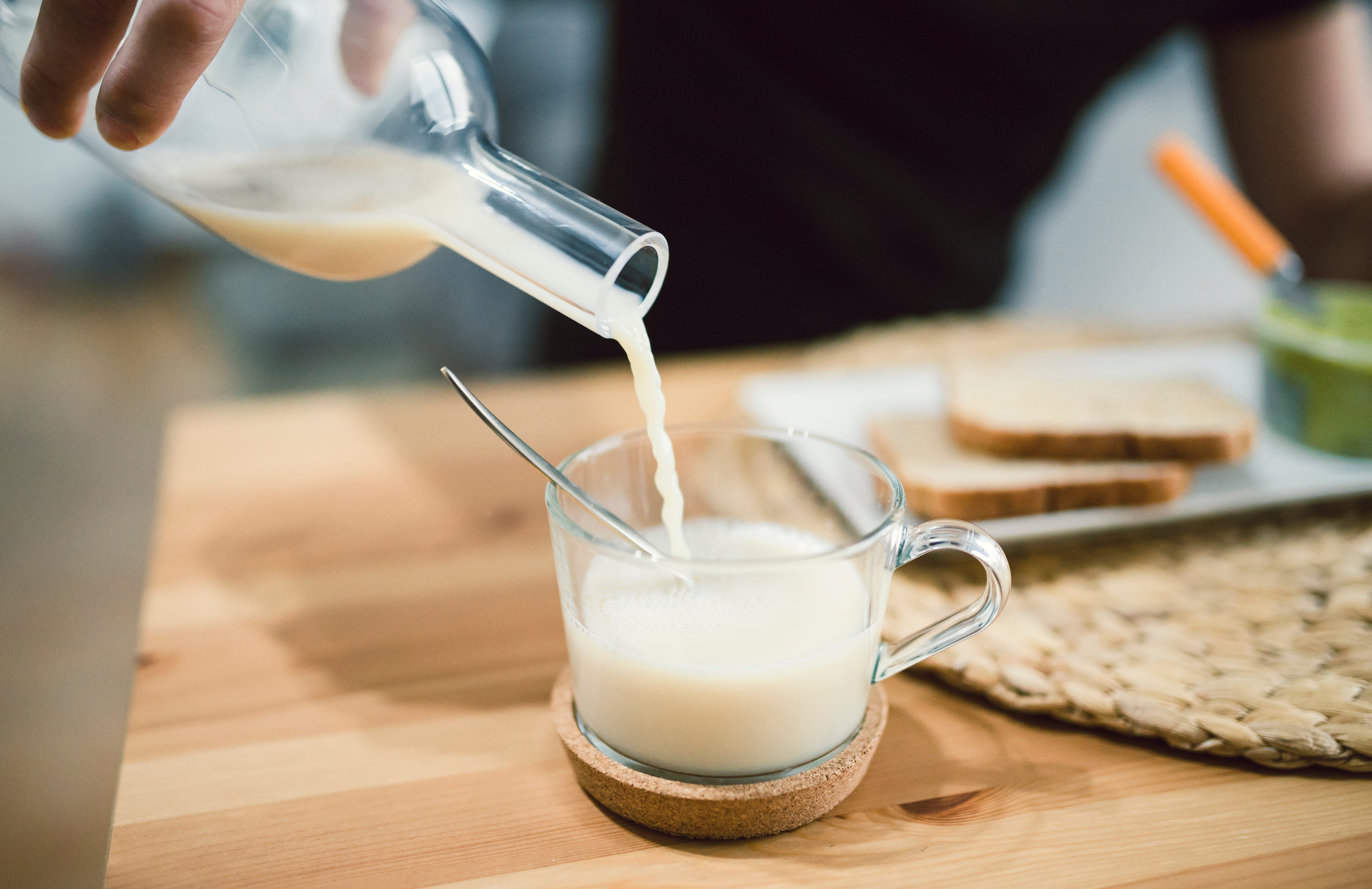 Pouring milk into a glass mug on a wooden table, with toast and a straw mat in the background