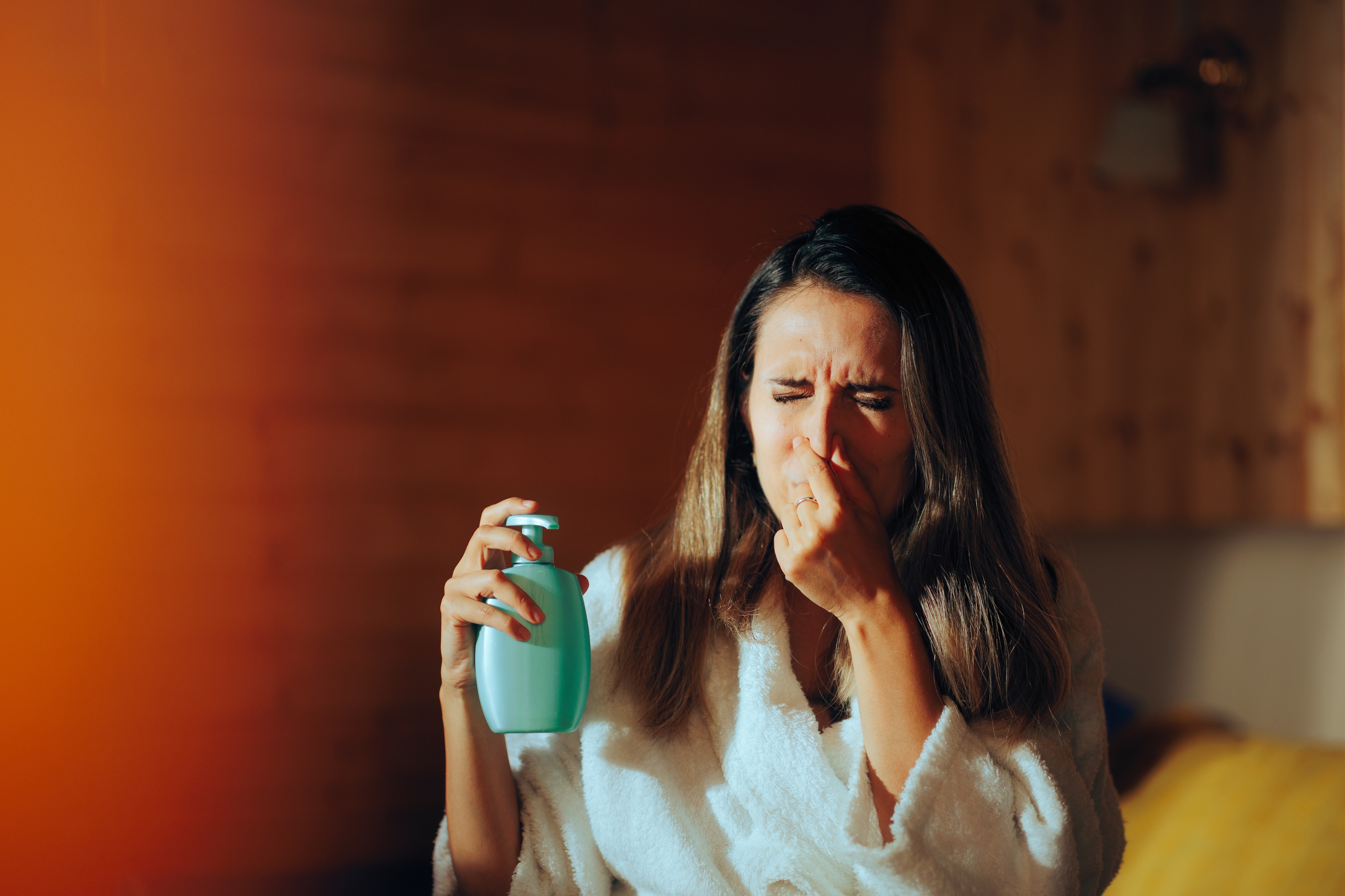 Person i en badekåpe nysing, holder en pumpeflaske, muligens reagerer på en duft eller et produkt