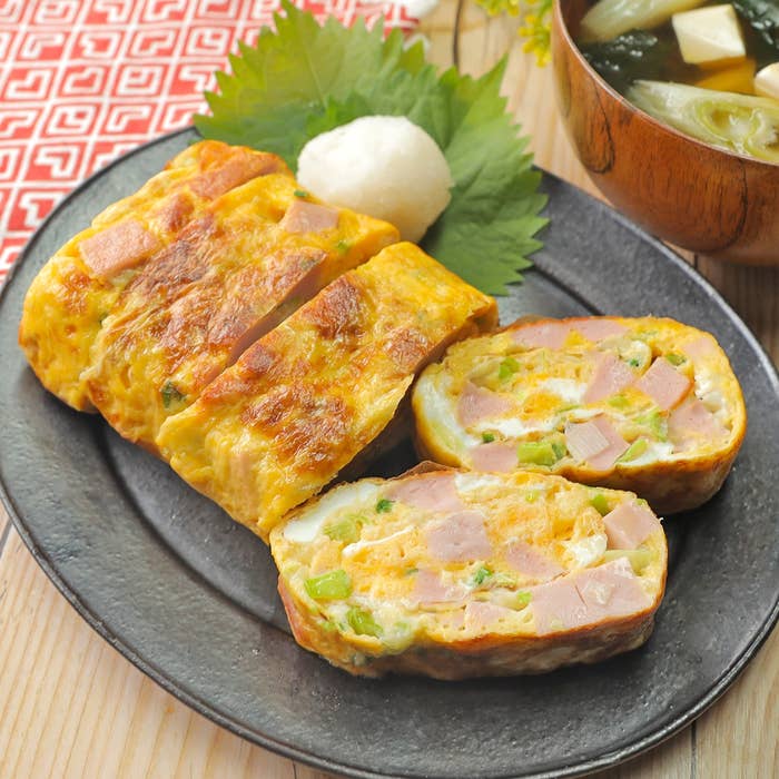 厚焼き卵にネギが入った料理が黒い皿に盛られている。隣にはおろし大根と和風スープが添えられている。