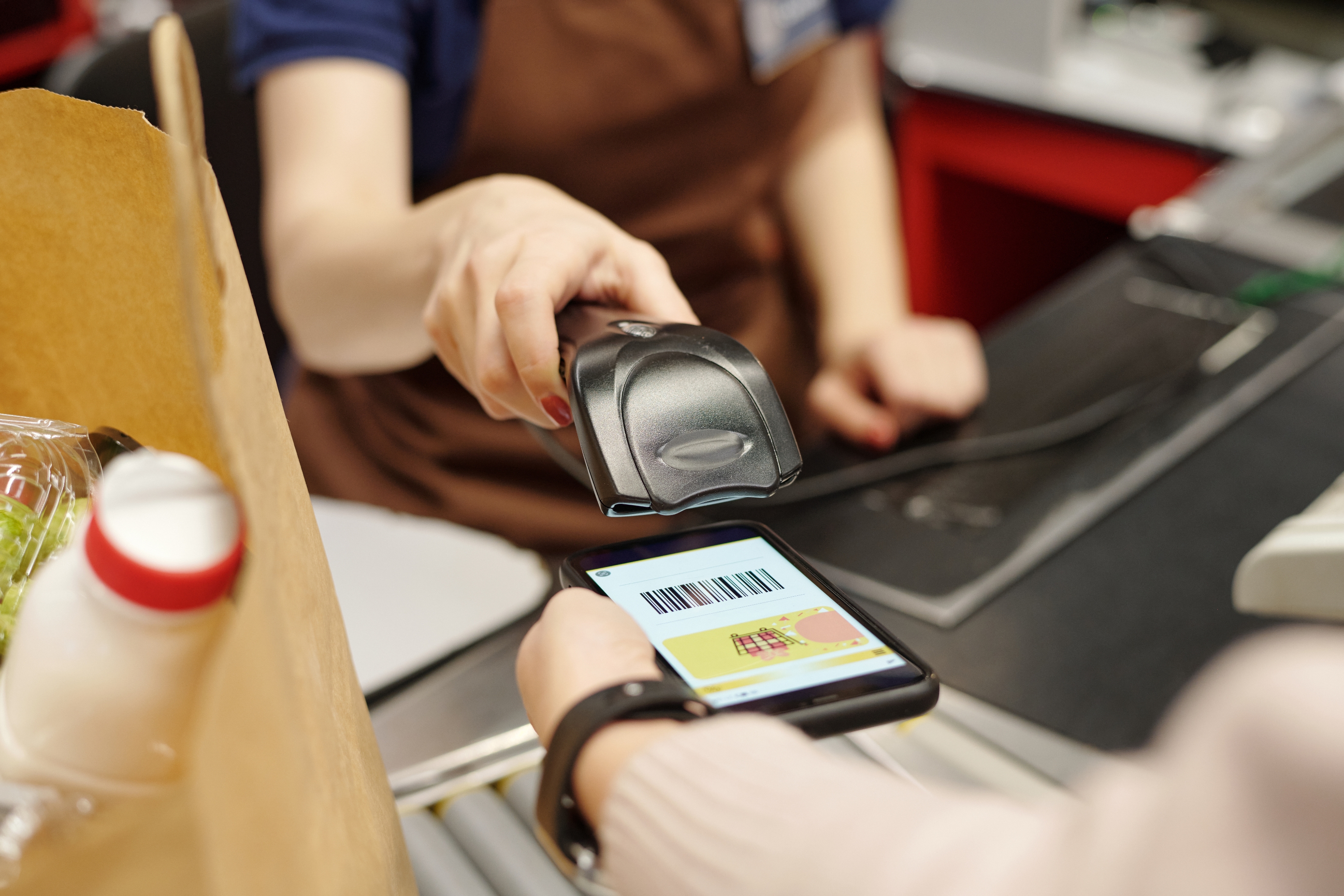 Person skanner strekkode på telefon i kassen, og holder dagligvarepose. Kasserer bruker håndholdt skanner