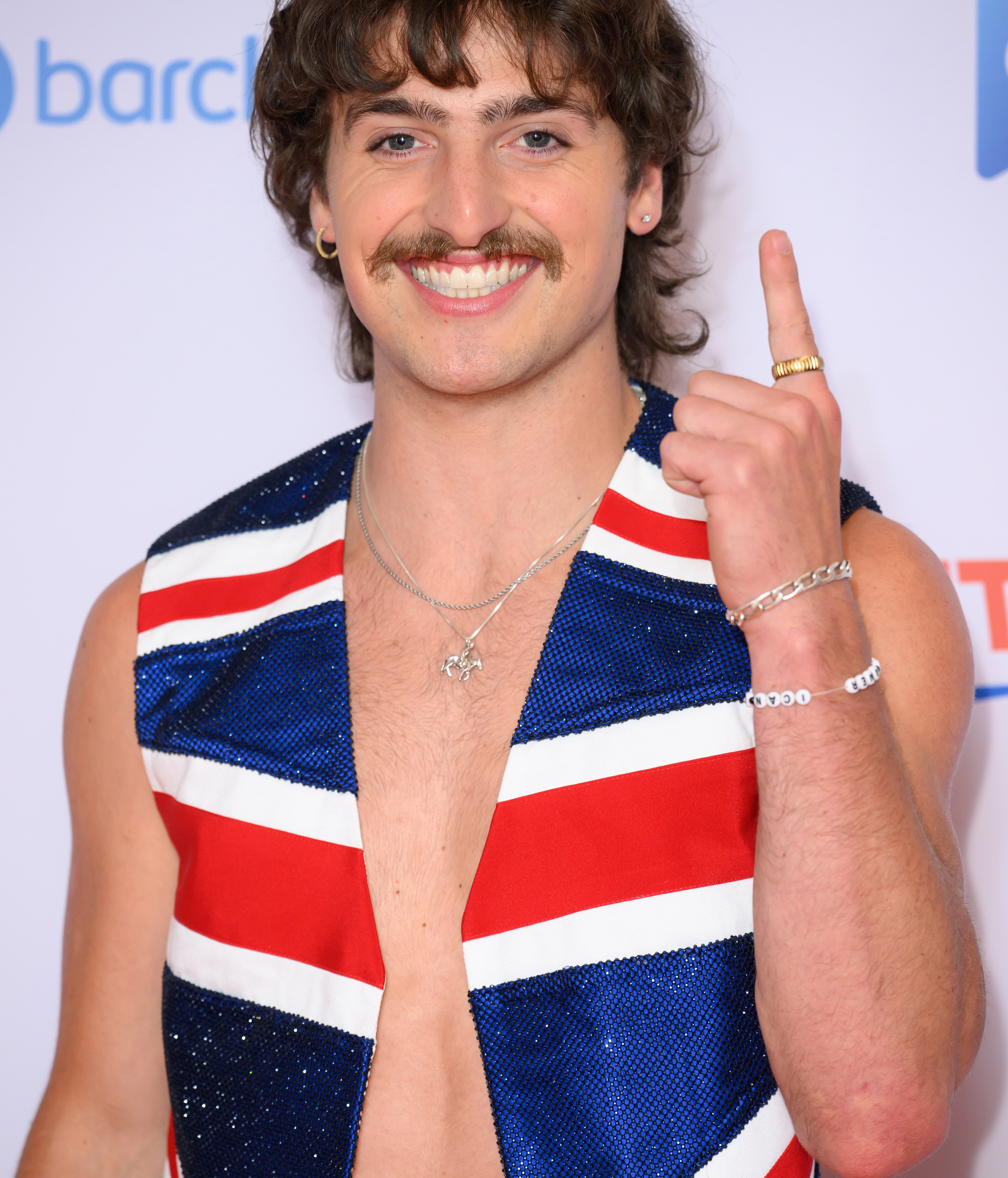 Personen smiler på den røde løperen iført et ermeløst antrekk med en Union Jack -design, tilbehør med et halskjede og armbånd