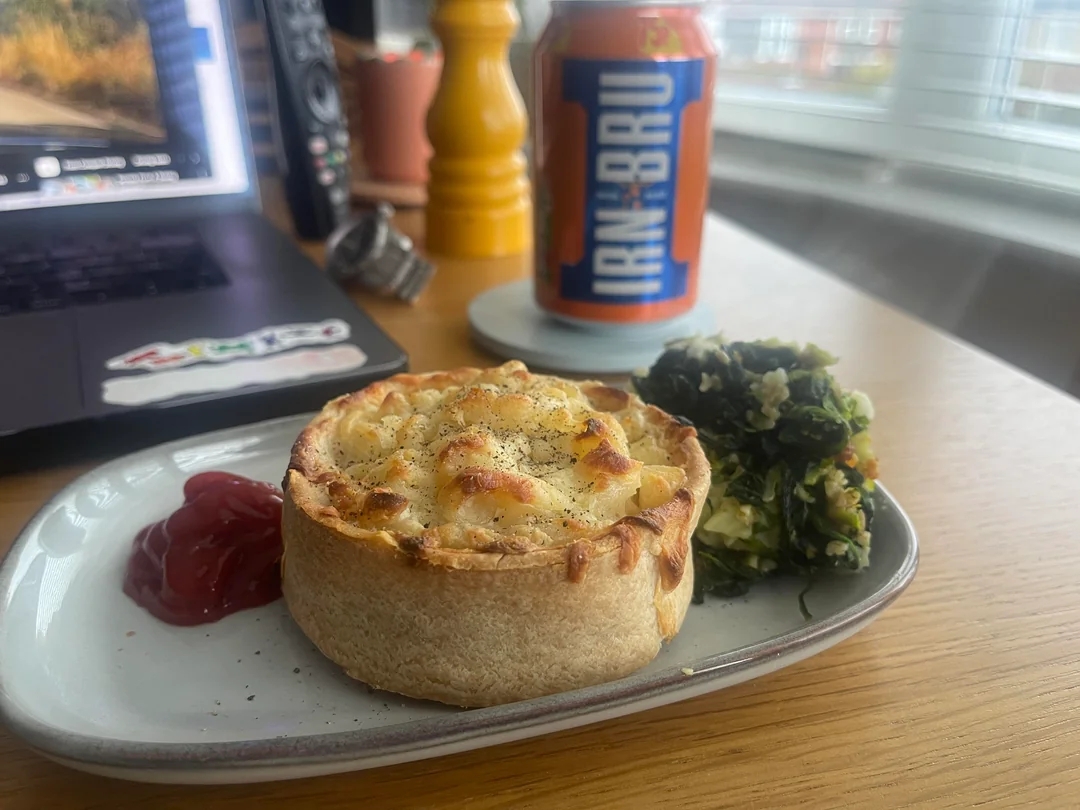 A savory pie with vegetables and ketchup on a plate, next to a can of Irn-Bru on a wooden table near a laptop