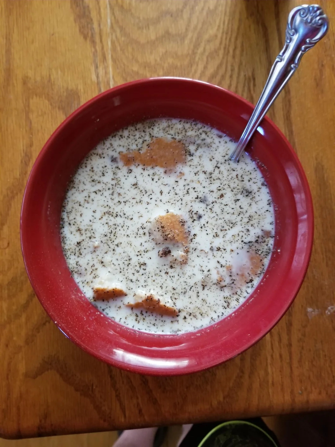 Bowl of milk with crumbled bread pieces and sprinkled herbs, placed on a wooden table, accompanied by a spoon