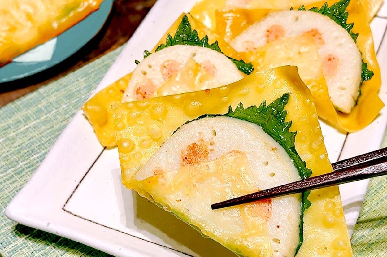 餃子の皮で巻かれたレンコンとエビの料理が皿に盛られている。箸で一つを持ち上げている。