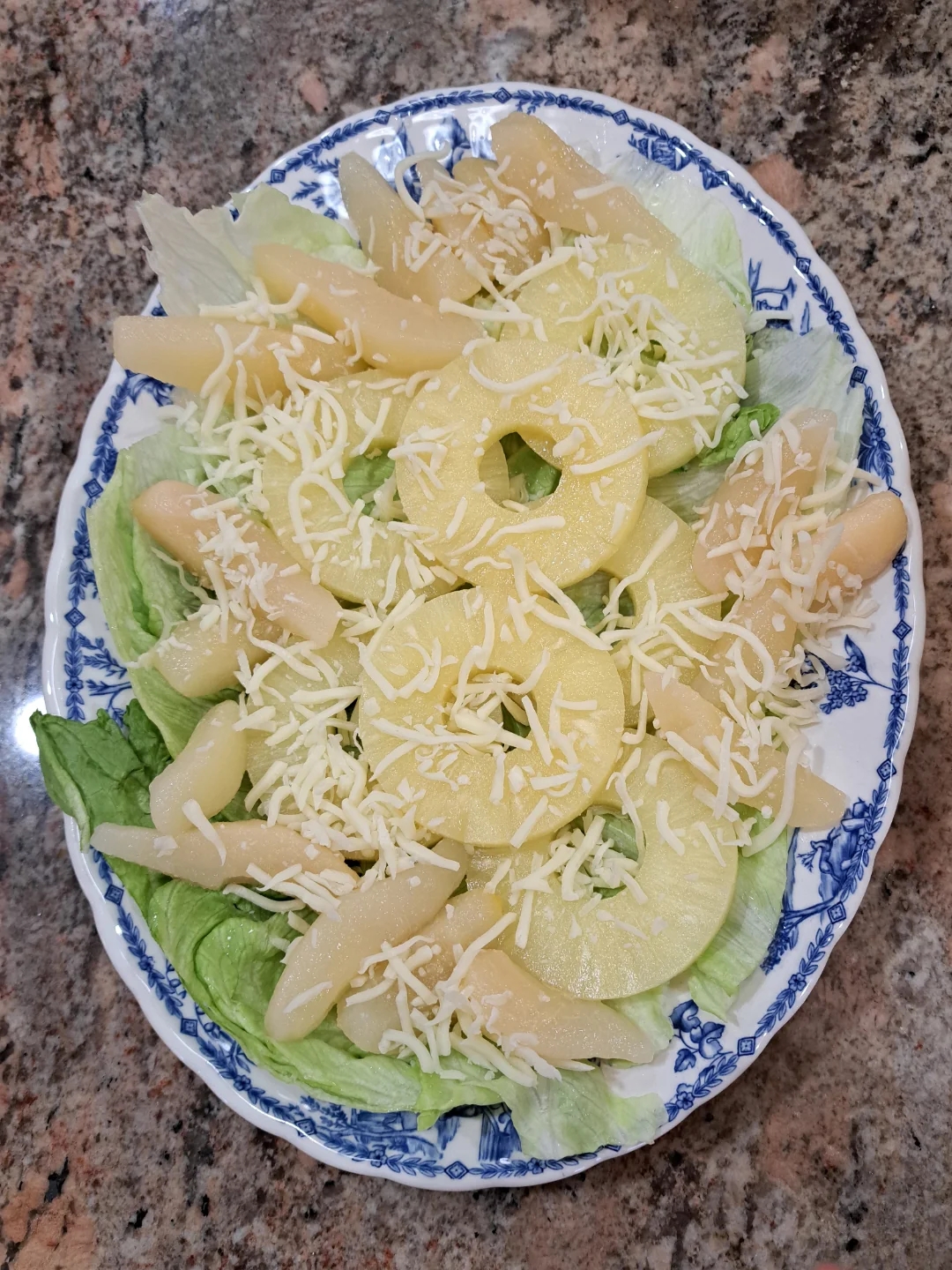 Salad with lettuce, pineapple rings, and white cheese on a blue-patterned plate, set on a stone surface