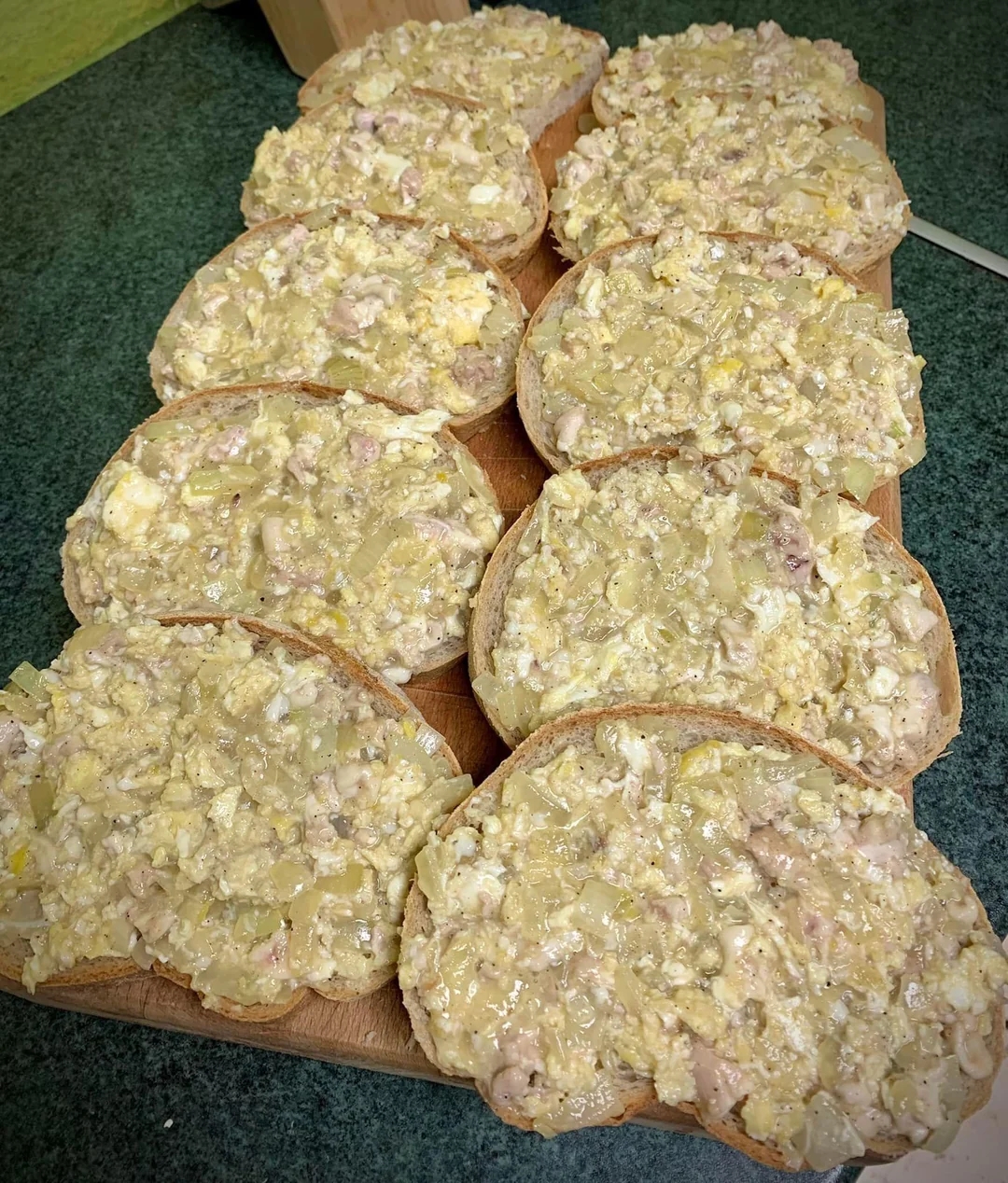 Open-faced sandwiches on a wooden board topped with a creamy mixture of chopped eggs, onions, and other ingredients