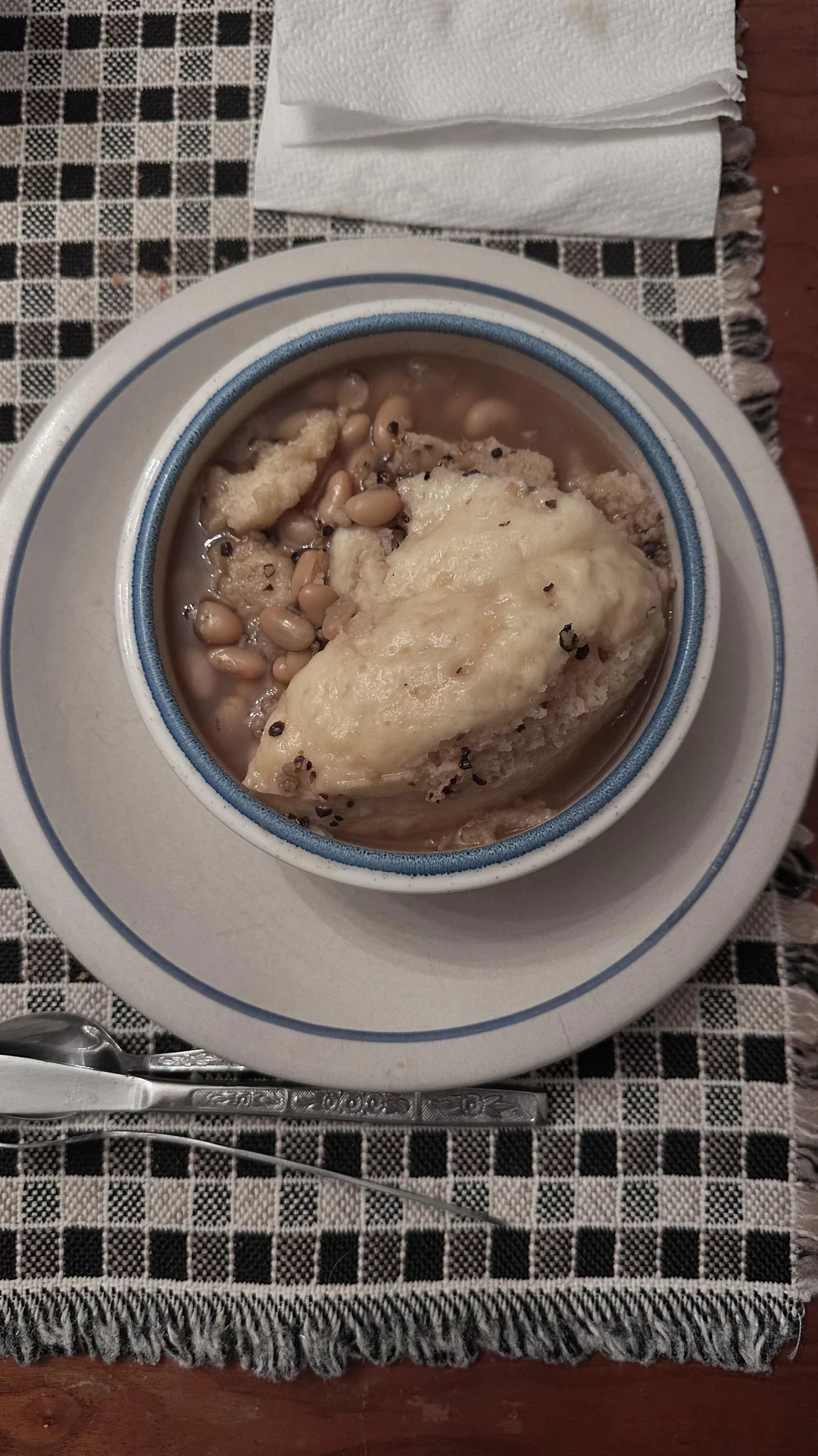 Beans with dumplings in a bowl on a checkered placemat, served with utensils and napkins
