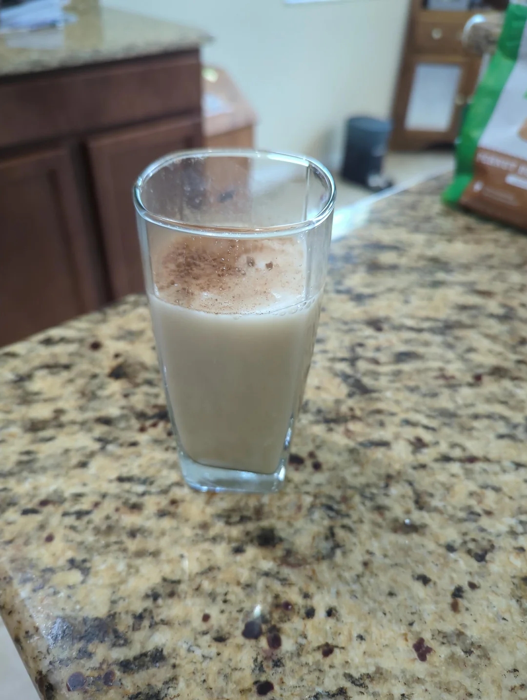 A tall glass of a creamy, spiced beverage on a granite countertop, with a sprinkle of cinnamon on top