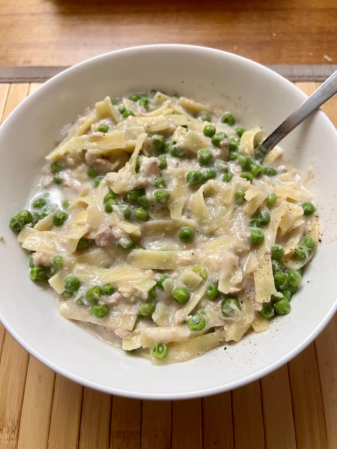 Bowl of creamy pasta with peas and chunks of meat, served with a spoon on a bamboo mat