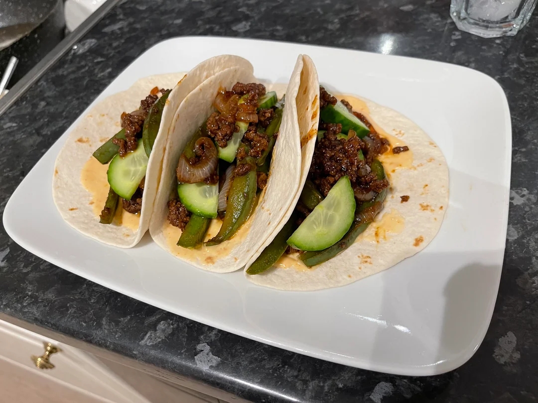 Three soft tacos filled with seasoned ground meat, sliced cucumbers, and bell peppers on a white plate