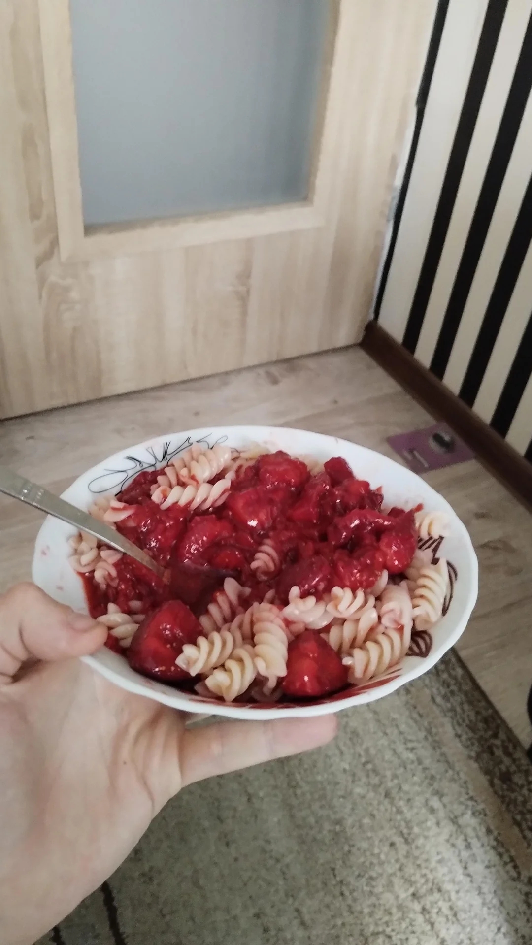 A plate of pasta with a bright red sauce, held in a hand over a patterned floor, near a door and striped wall