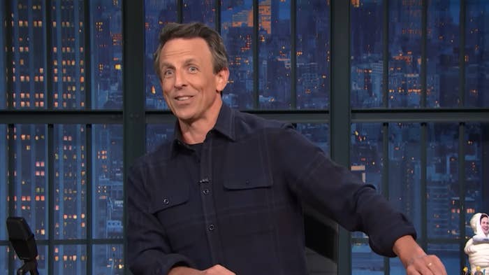 Man in casual shirt appears on what looks like a talk show set, smiling slightly, with a cityscape visible through large windows behind him