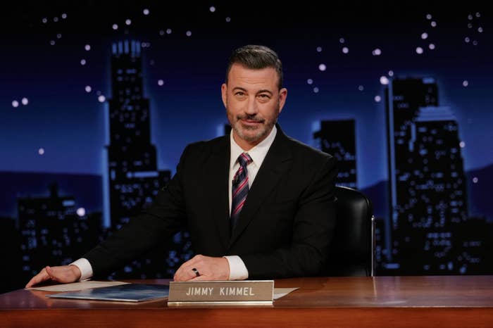 Person in a suit sits at a talk show desk with a nighttime cityscape backdrop