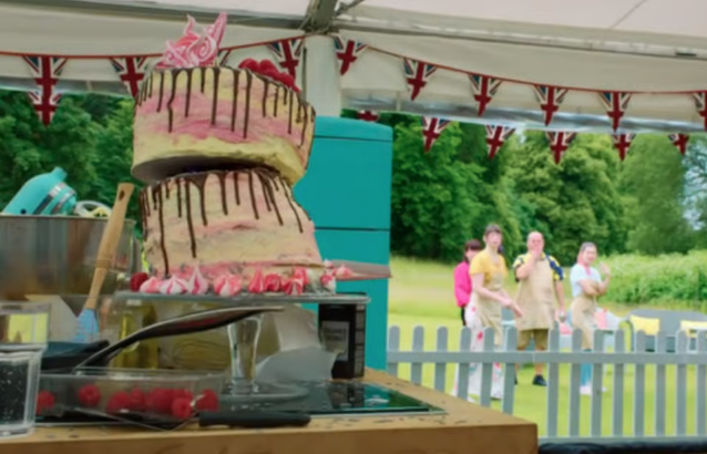 Layered cake with chocolate drizzle on display in a tent with people in the background and Union Jack bunting overhead