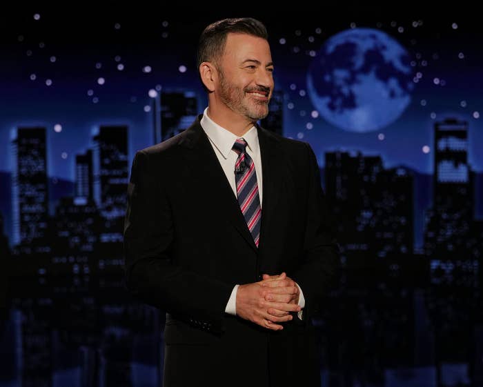 A man is smiling on a nighttime TV set, wearing a dark suit and striped tie. Cityscape and large moon visible in the background