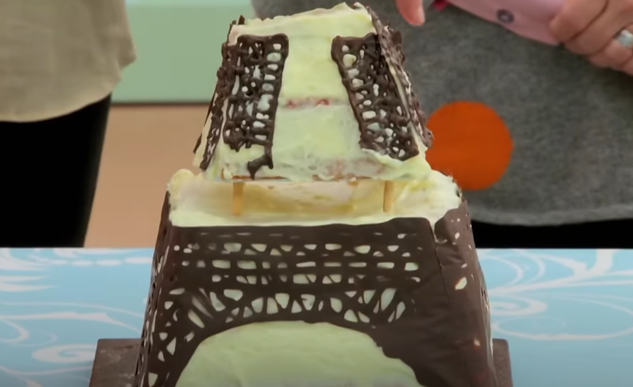 Cake resembling the Eiffel Tower, detailed with chocolate latticework, with visible frosting and partial structure collapse at the top