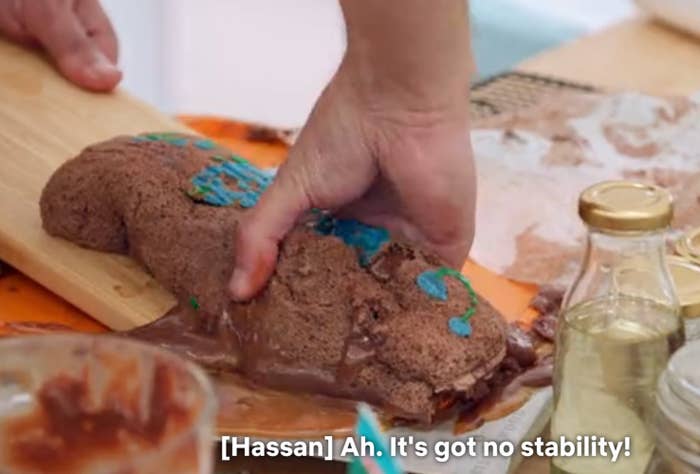 A person grips a partially collapsed chocolate cake as melted chocolate oozes out. Subtitles read: &quot;[Hassan] Ah. It&#x27;s got no stability!&quot;
