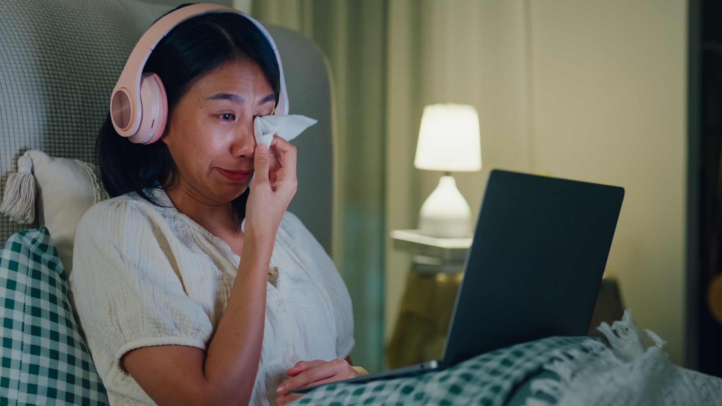 Kvinne iført hodetelefoner og pyjamas sitter i sengen og tørker tårer mens du ser på noe på en bærbar datamaskin