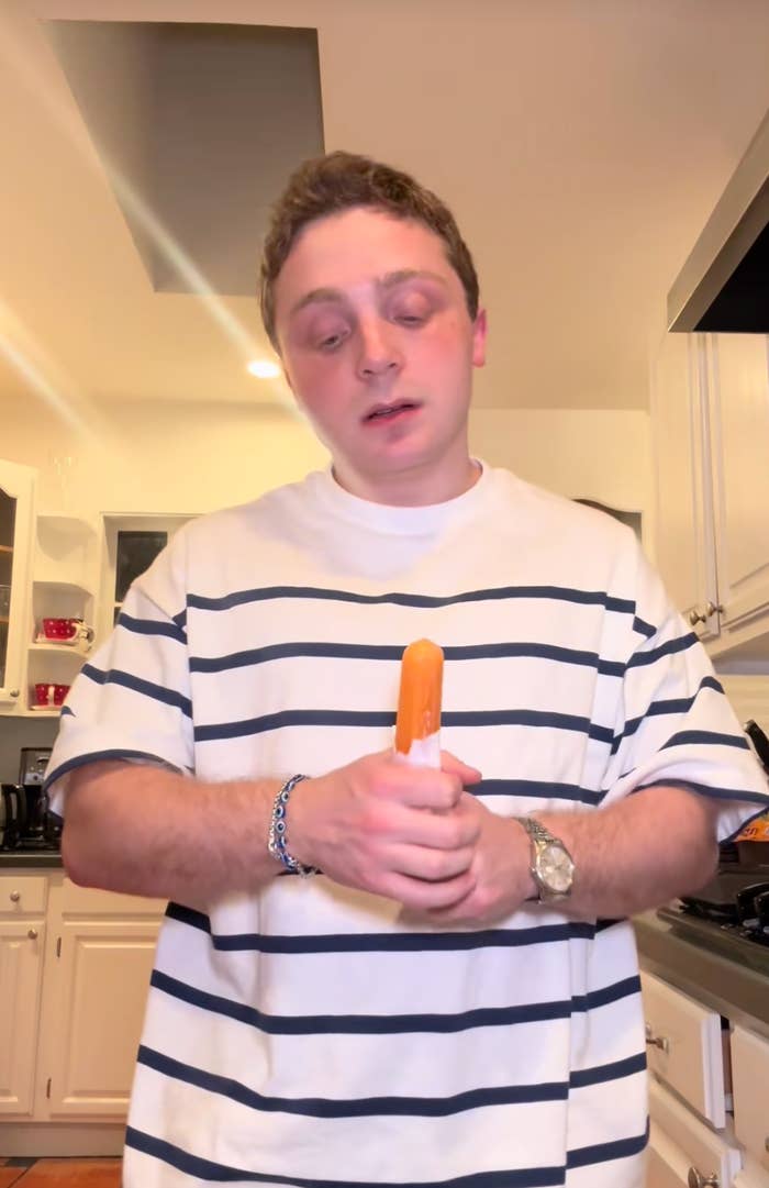 A person in a kitchen holding a popsicle, wearing a striped t-shirt and bracelet