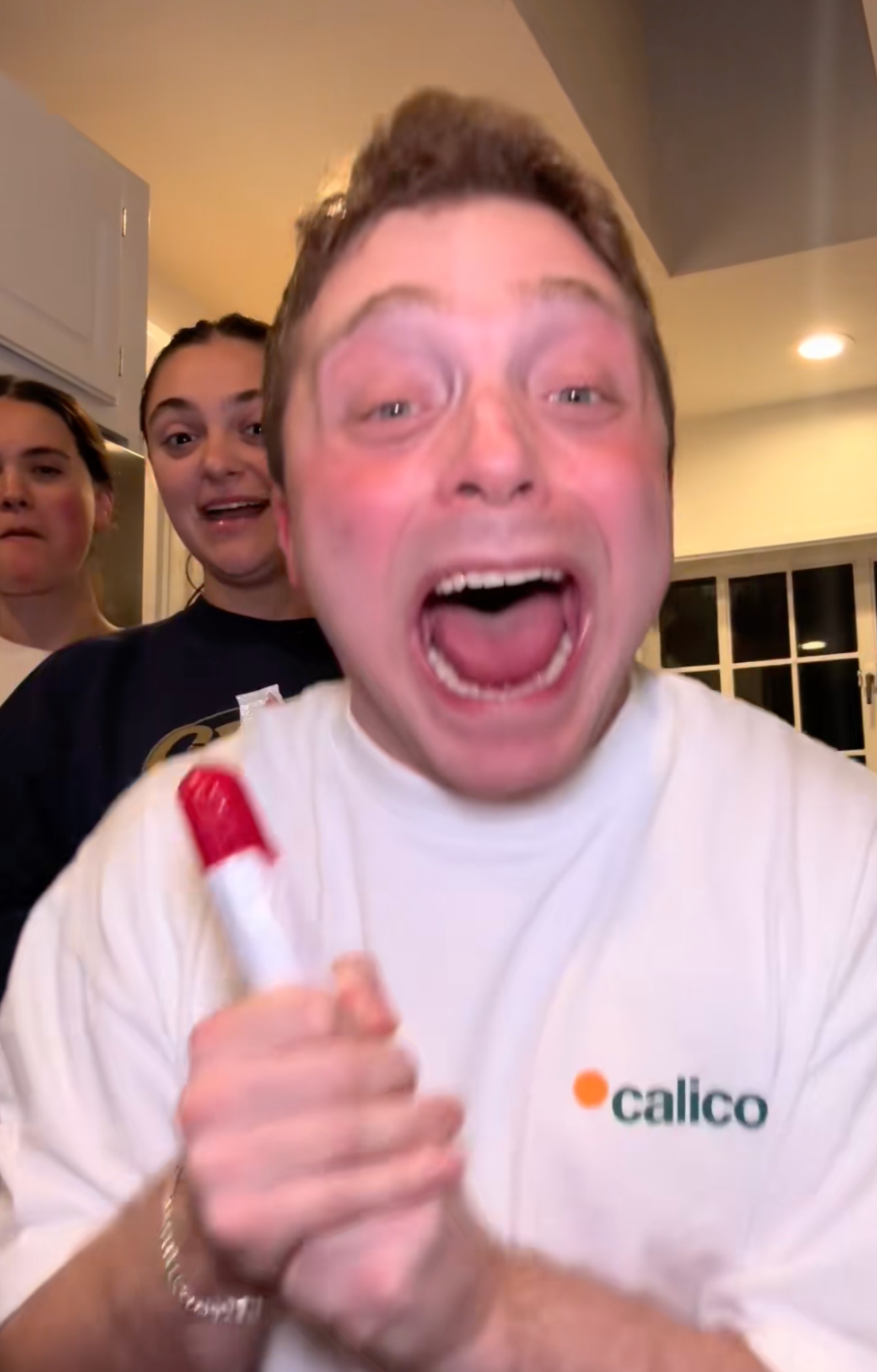 A person with an excited expression holds a tube-like object while standing with two others in a kitchen