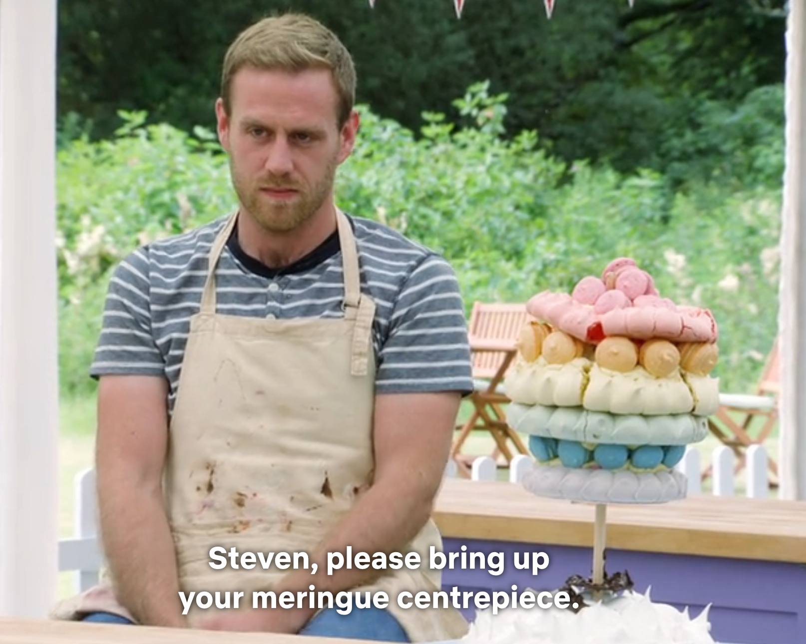 A contestant in a striped shirt and apron stands next to a colorful meringue centerpiece on a baking show set outdoors
