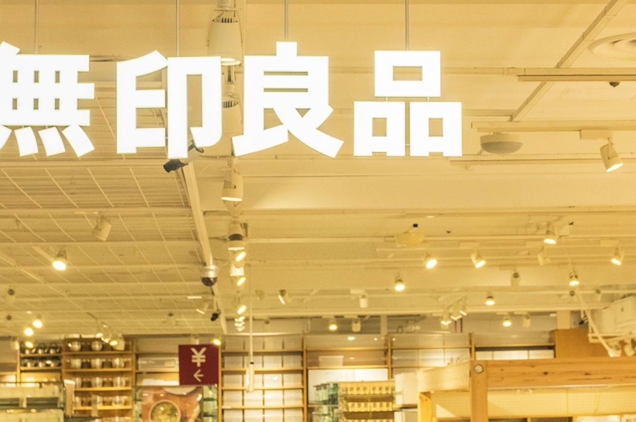 A spacious store with home goods and clothing, featuring organized shelves and signs with Japanese text. Shoppers are browsing the aisles