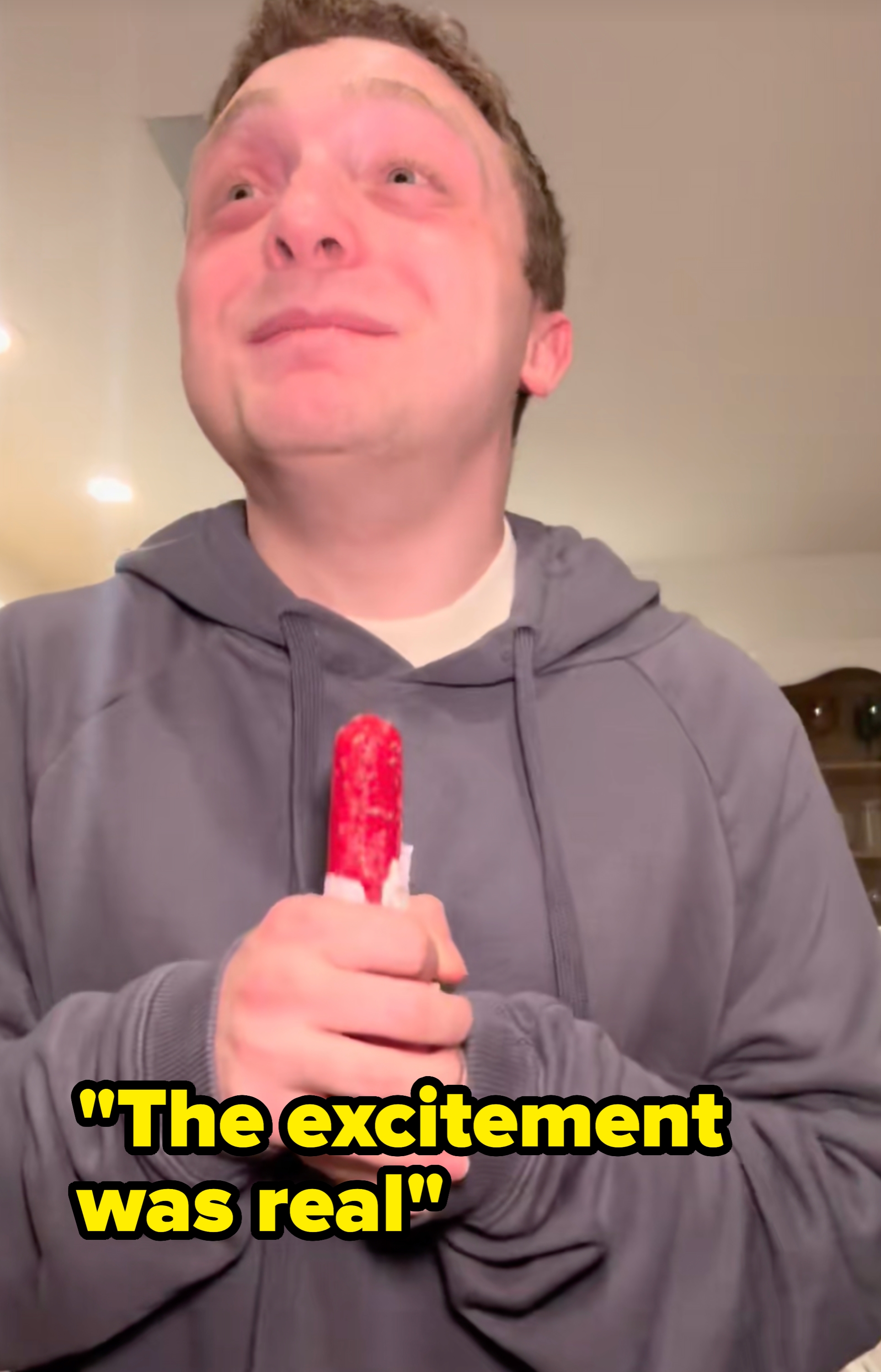 Person in a kitchen holding a popsicle, wearing a T-shirt with an artistic print. Another person is partially visible behind them, smiling