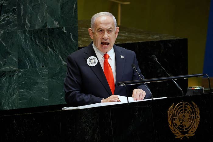 Person speaking at a United Nations podium, wearing a suit with an orange tie and a pin, appearing to address an audience