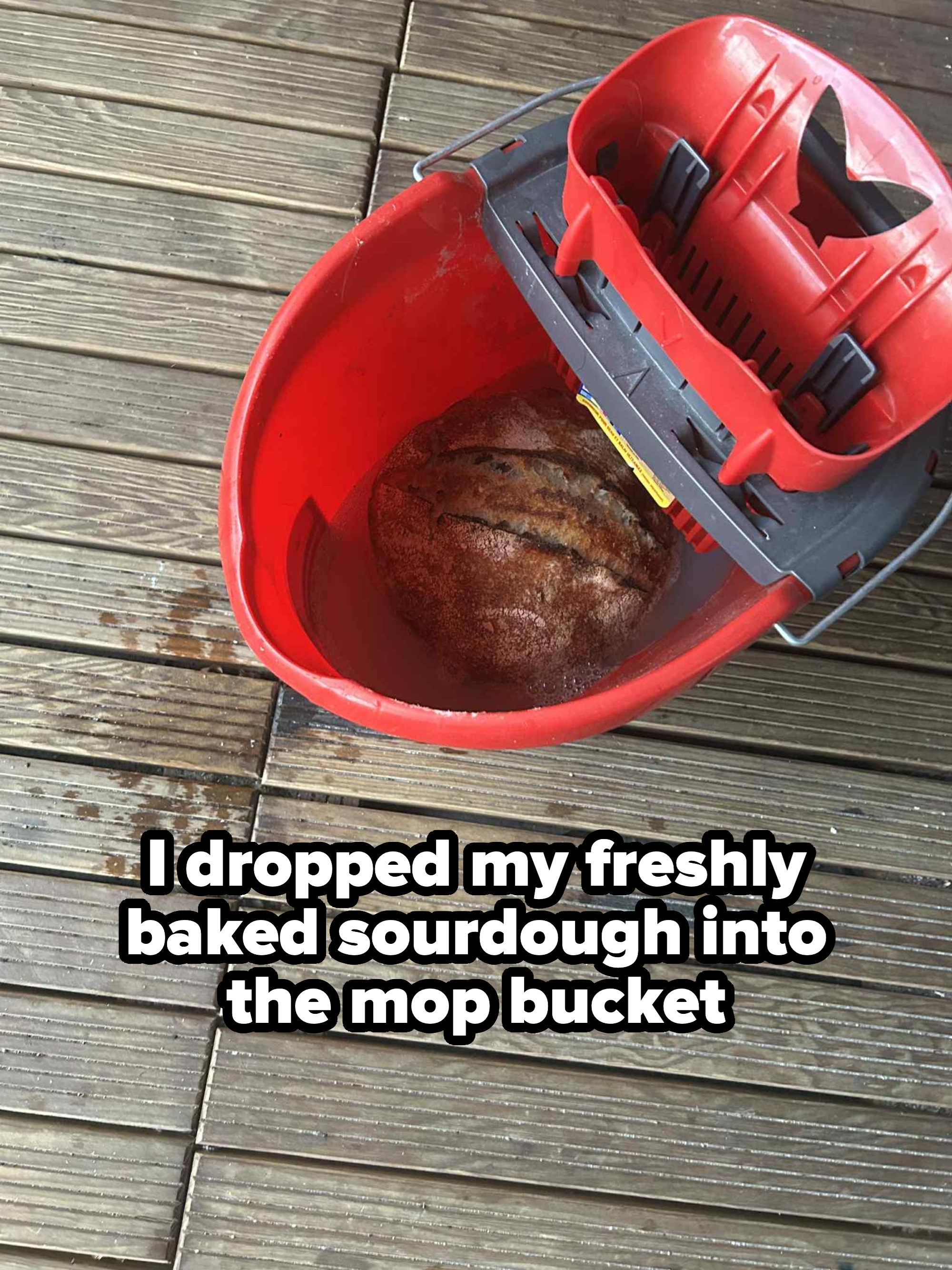 A loaf of bread is humorously placed inside a red mop bucket on a wooden floor