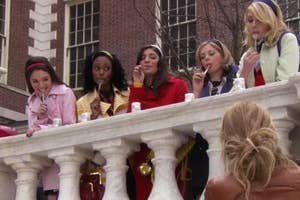 Group of stylishly dressed girls in "Gossip Girl" stand at a railing, enjoying lollipops and looking smug, high school behind them