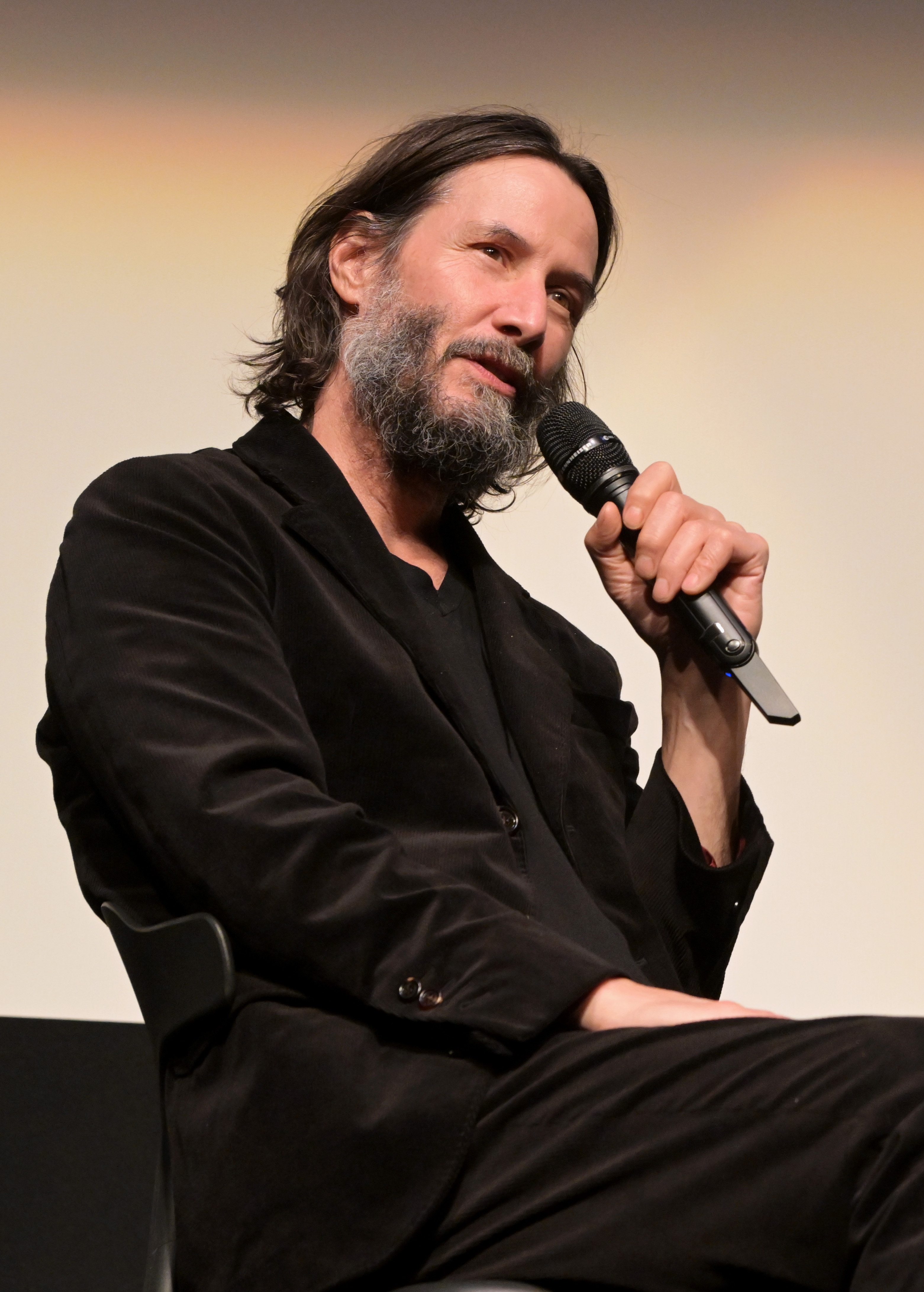 Keanu Reeves in a suit sits, holding a microphone, speaking at an event