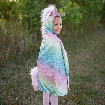 Child wearing a sparkling unicorn costume with a hood, ears, and tail, lasting outdoors and smiling astatine the camera