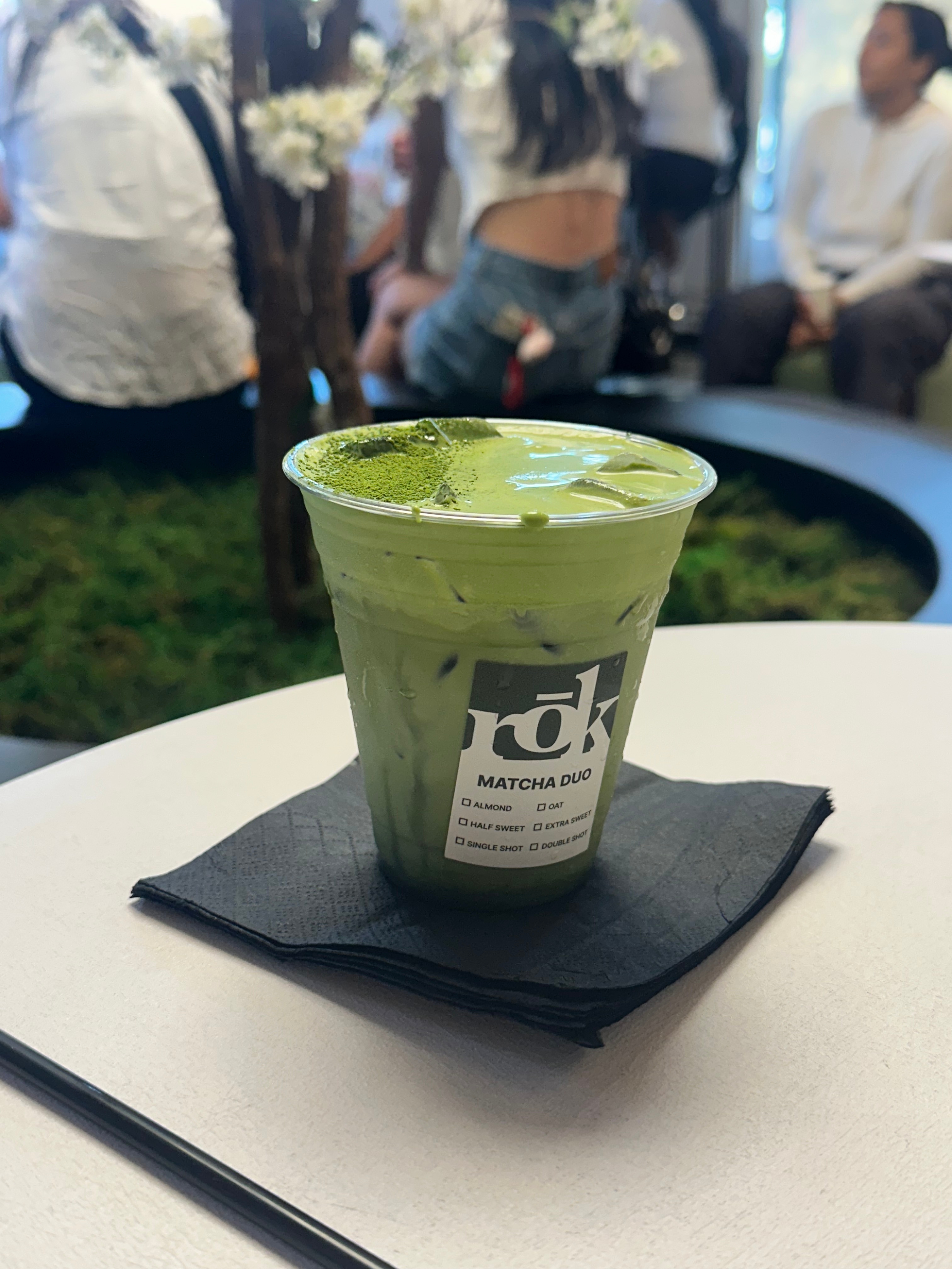 Iced matcha latte in a clear cup on a black napkin, with seated people in a blurred background