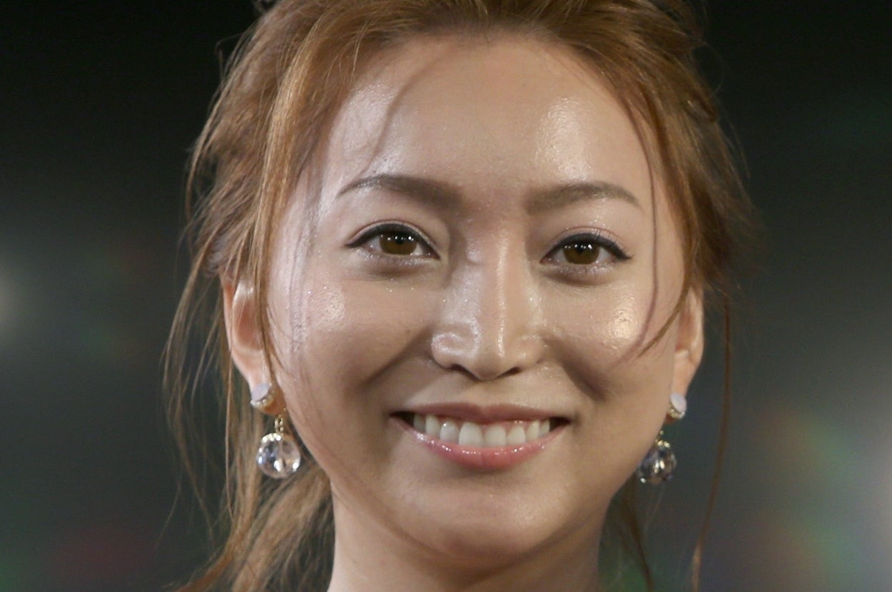 女性がカメラに微笑みながら立っている。髪はアップスタイルで、大ぶりのイヤリングを着けている。