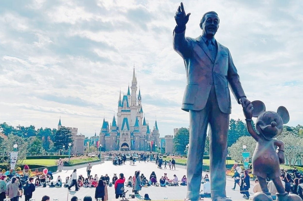 ディズニーランドのシンデレラ城前で、ウォルト・ディズニーとミッキーマウスの像が観光客で賑わう公園に立っている。