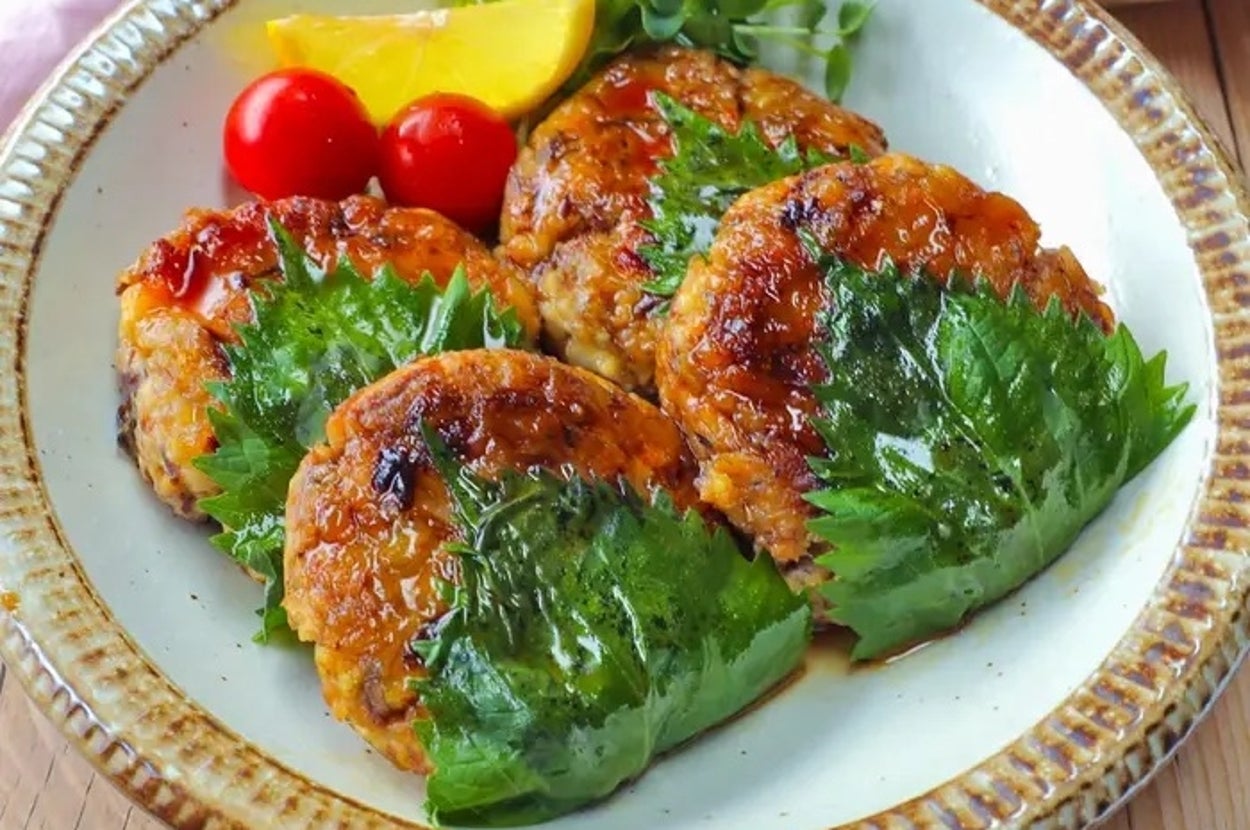 大皿に盛られた鶏つくねハンバーグが3つ、大葉と一緒に並んでいる。隣にミニトマトとレモンも添えられている。