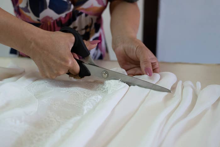 Hands use scissors to cut white fabric with lace detail on a table