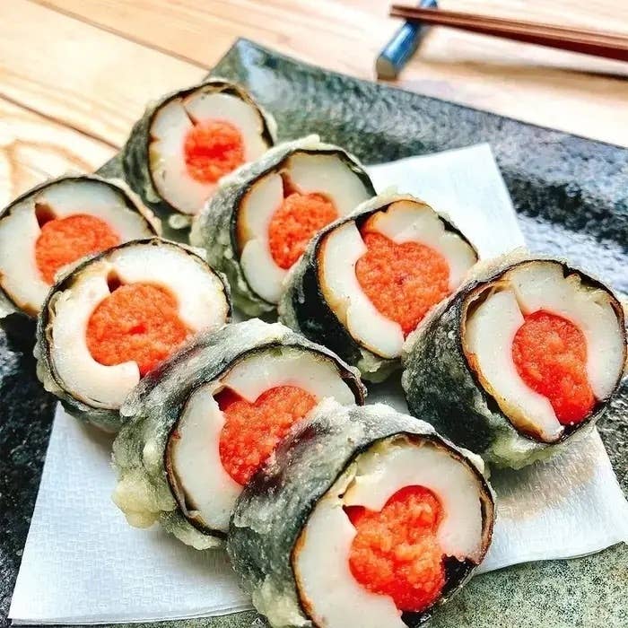 天ぷらで揚げられた巻寿司が黒い皿に並んでいる。内部にはオレンジ色の具材が見える。