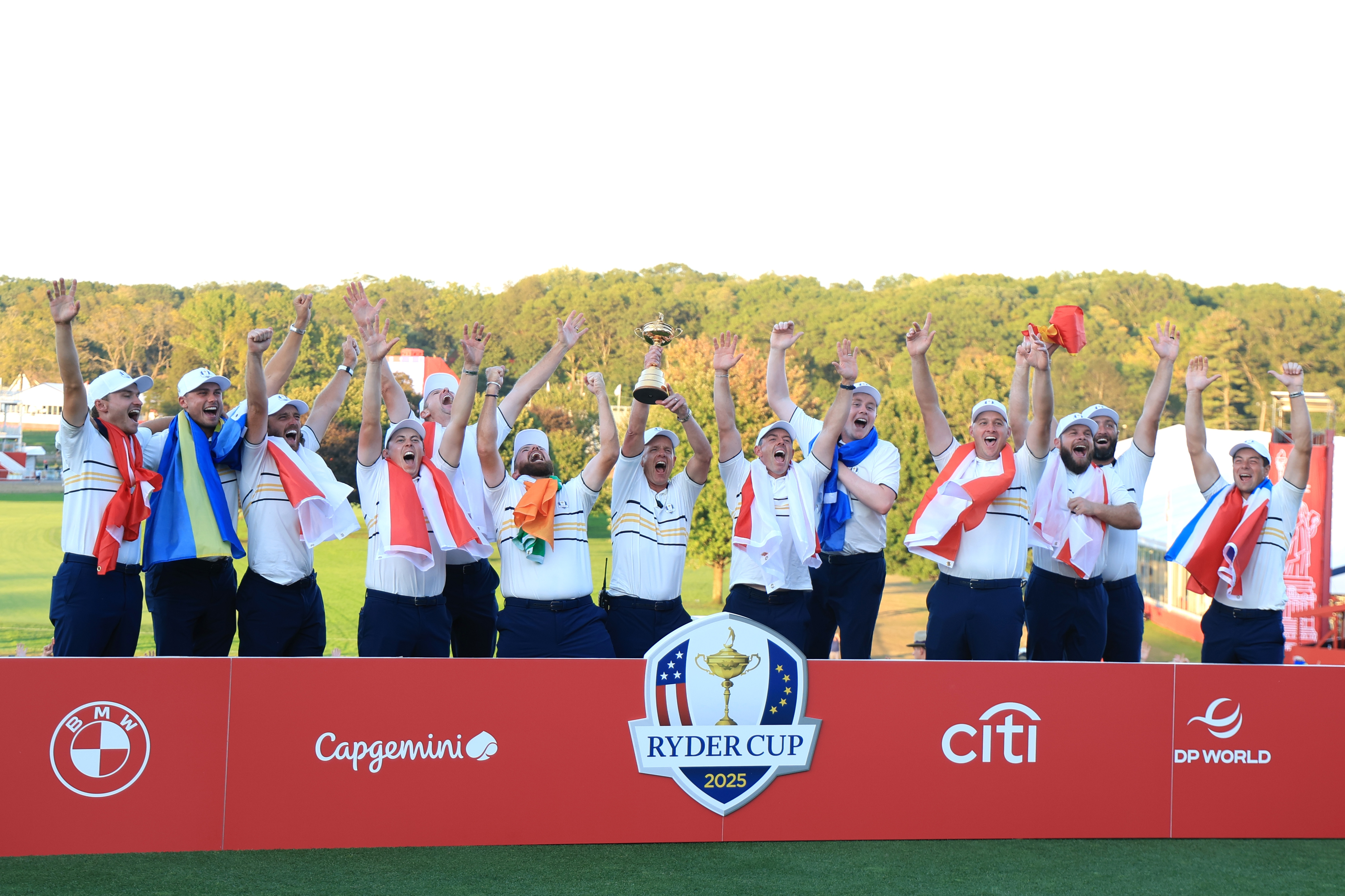 Golfers celebrate winning the Ryder Cup, holding a trophy and raising their arms on a stage with sponsor logos