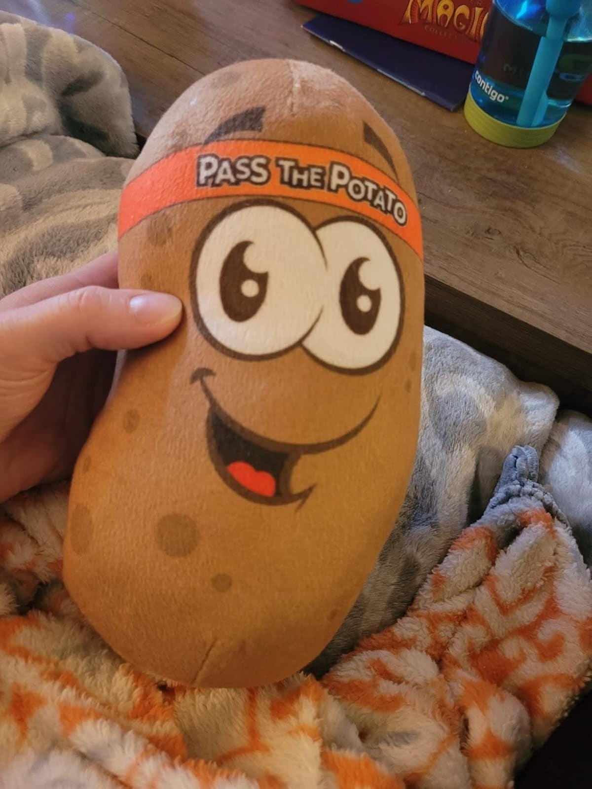 Hand holds a plush toy potato with cartoon eyes and a smile, labeled "Pass the Potato," on a cozy blanket